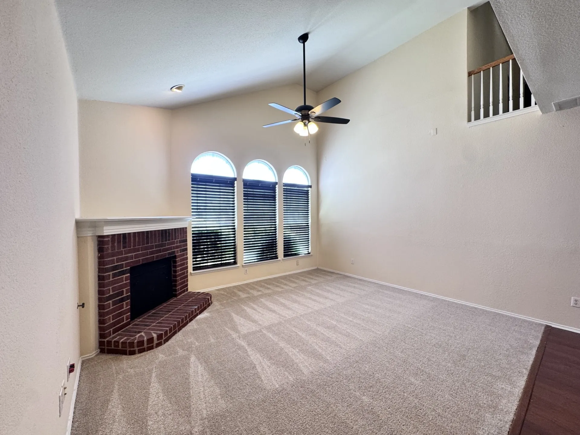 Room featuring a vaulted ceiling with a ceiling fan, a brick fireplace with a white mantel, three arched windows with blinds, and carpet flooring