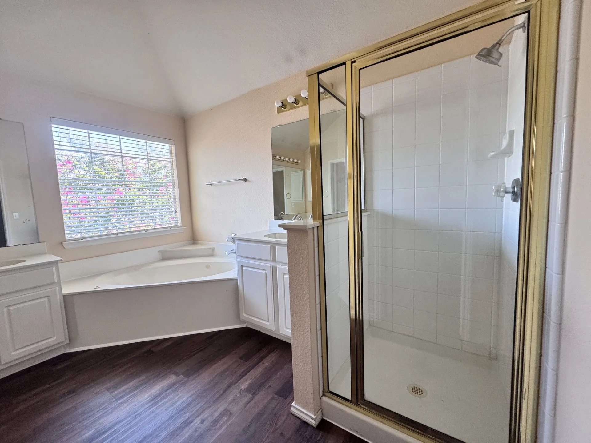 Soaking tub with a large window, dual vanities, wood-finish flooring, and a tiled shower with a glass enclosure