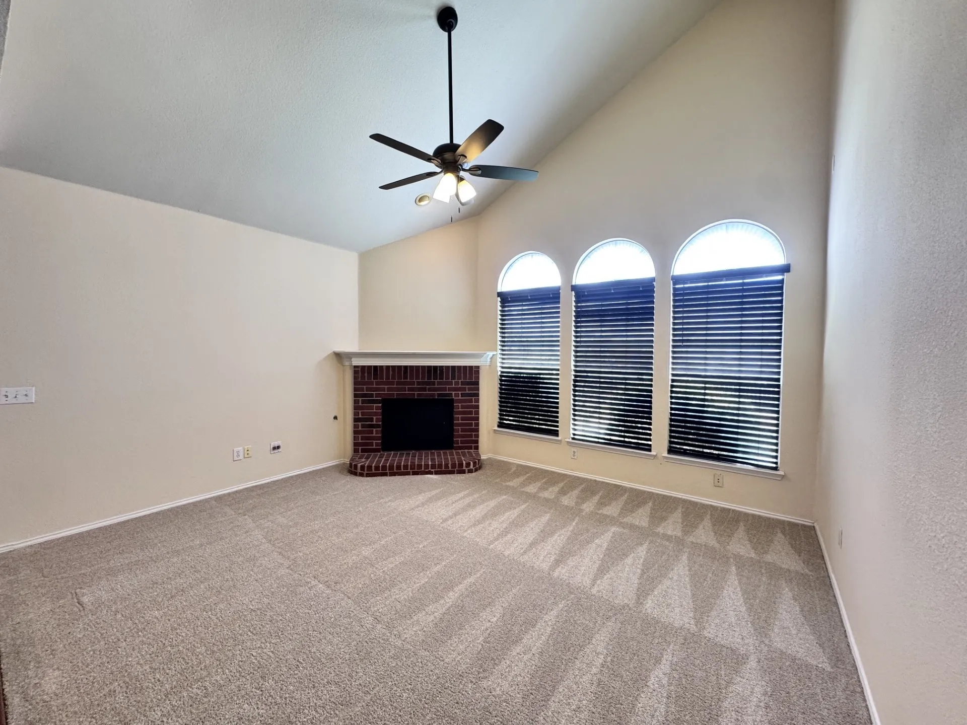 Vaulted ceiling with a ceiling fan, three arched windows, corner brick fireplace with a mantel, and neutral carpeting