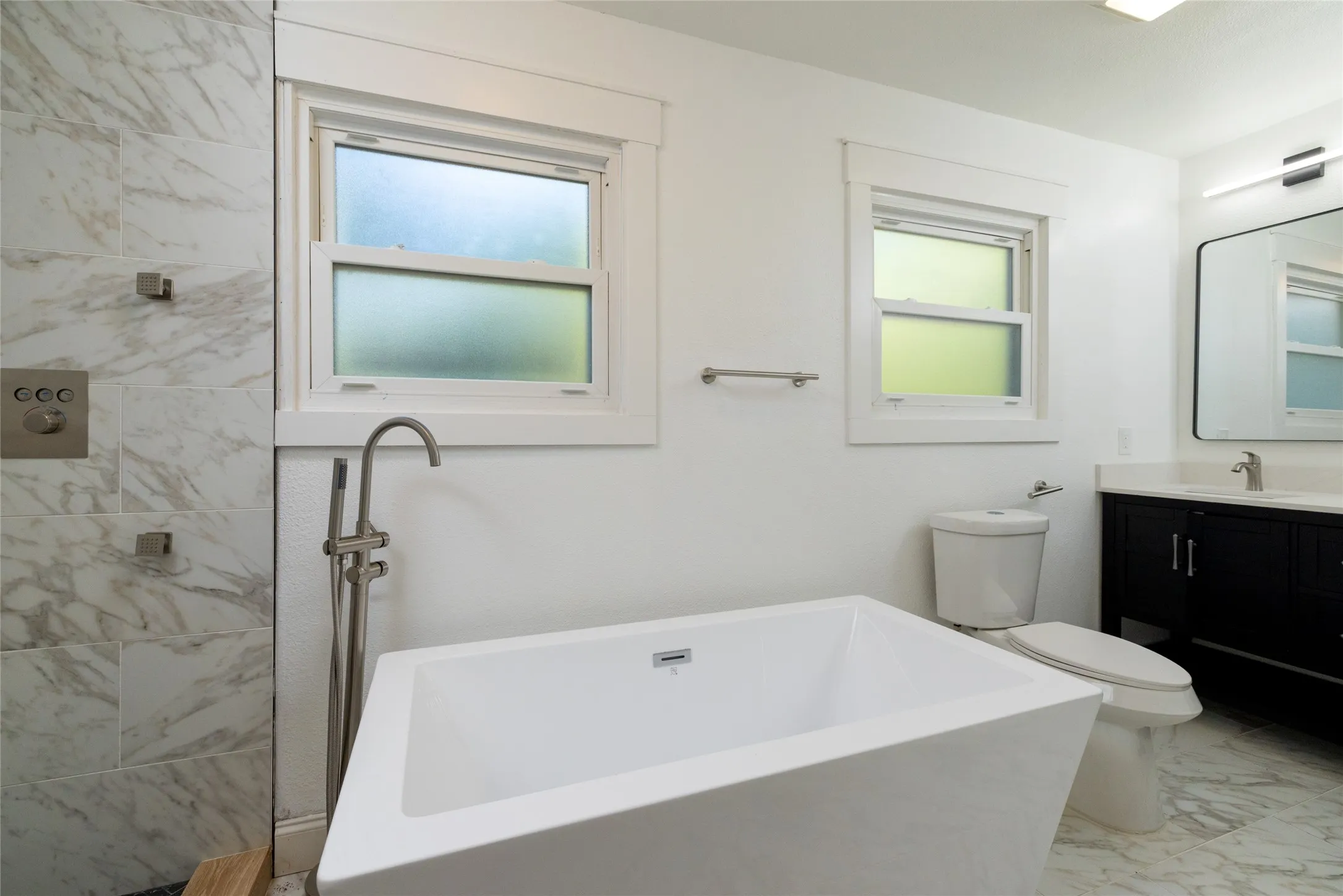 Freestanding soaking tub with floor-mounted brushed nickel faucet, large format tile shower, dual-pane windows, and a vanity with a rectangular sink and mirrored cabinet