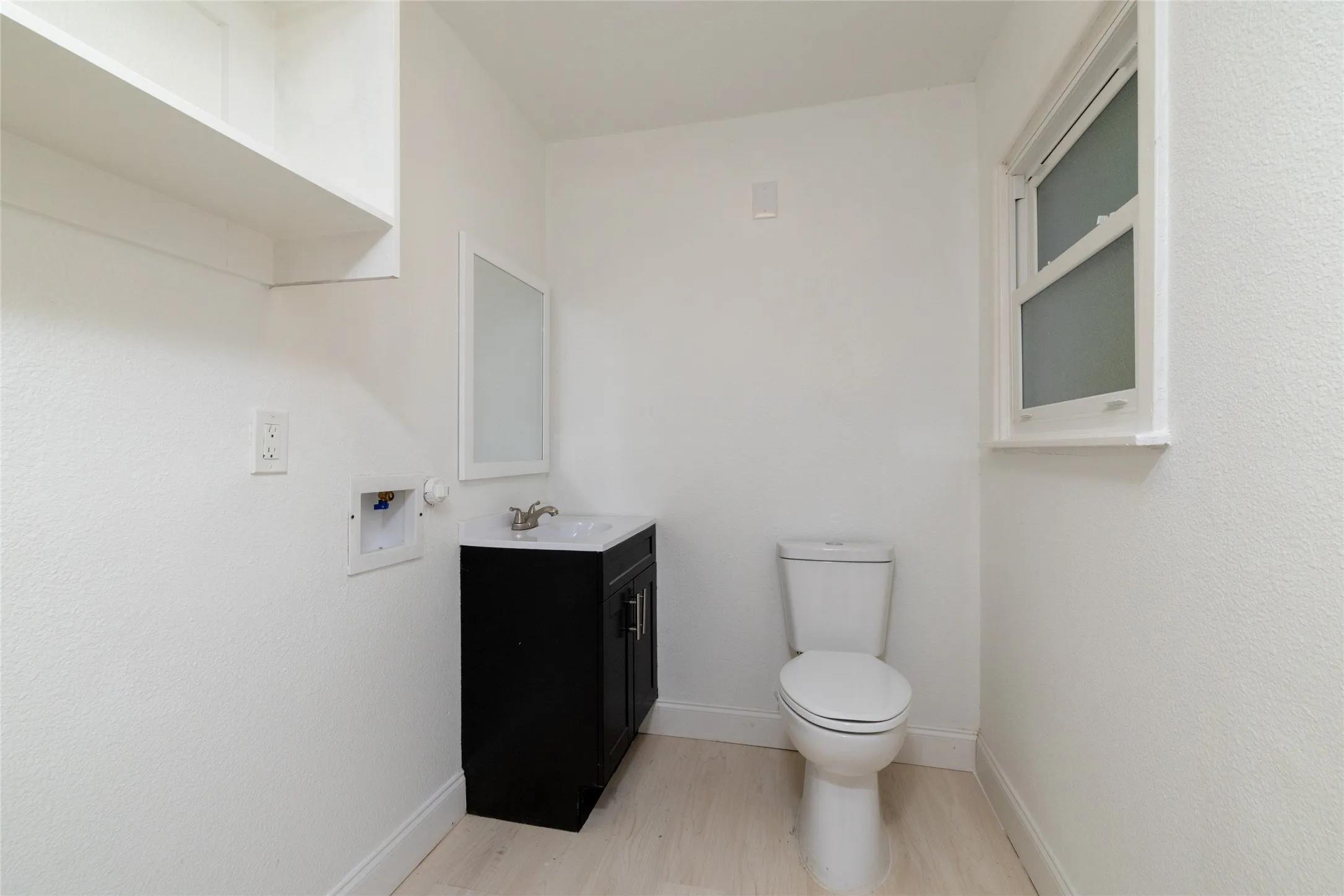 White wood-finish flooring, white walls, built-in shelving, dark vanity with integrated sink, and a single window with white trim