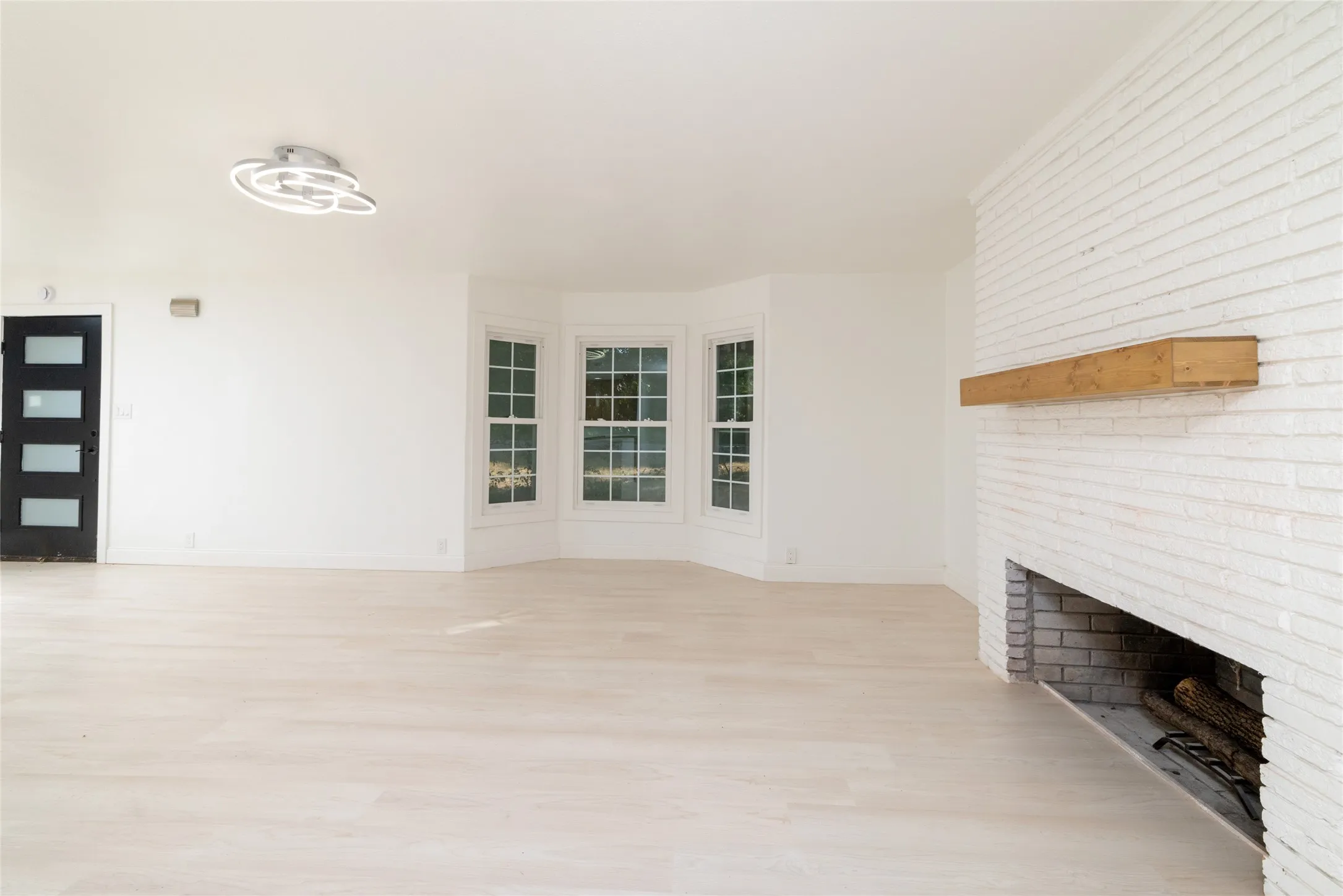 Whitewashed brick fireplace with a wood mantle, bay windows, contemporary ceiling light fixture, and light wood-finish flooring