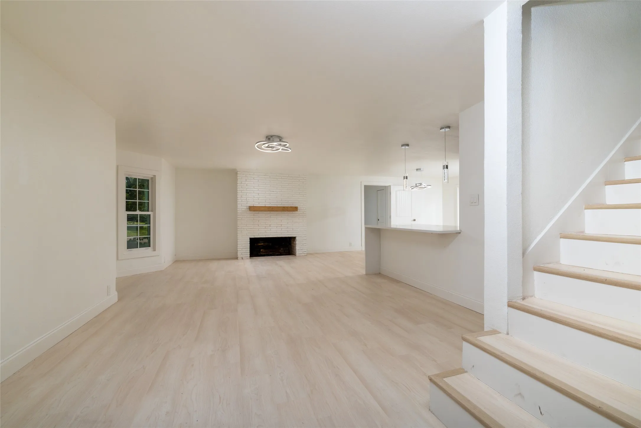 Light wood-finish flooring, a brick fireplace with a wood mantle, a kitchen pass-through counter, a staircase with wood treads, and contemporary pendant lighting