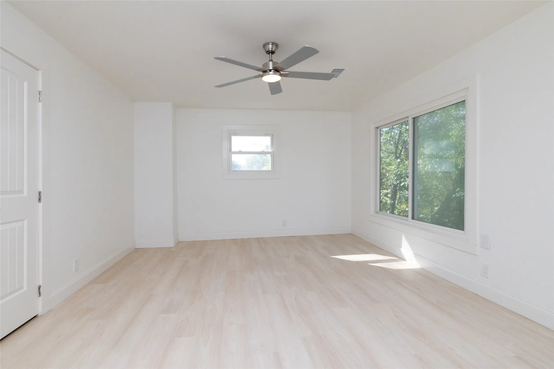Light wood-finish flooring, white walls, ceiling fan with integrated lighting, and multiple windows