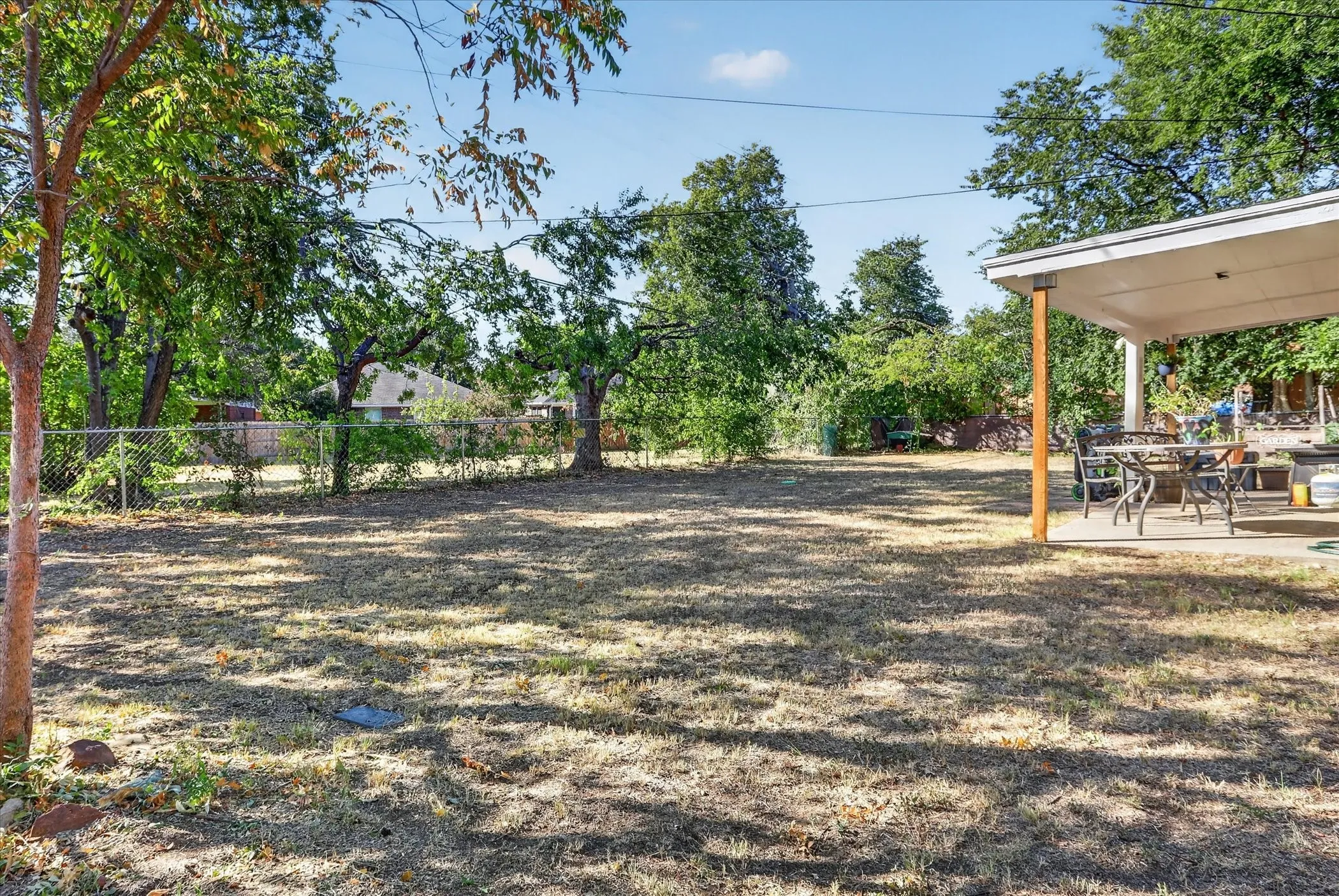 Expansive yard space featuring a chain-link fence, mature trees, and a covered patio with concrete flooring