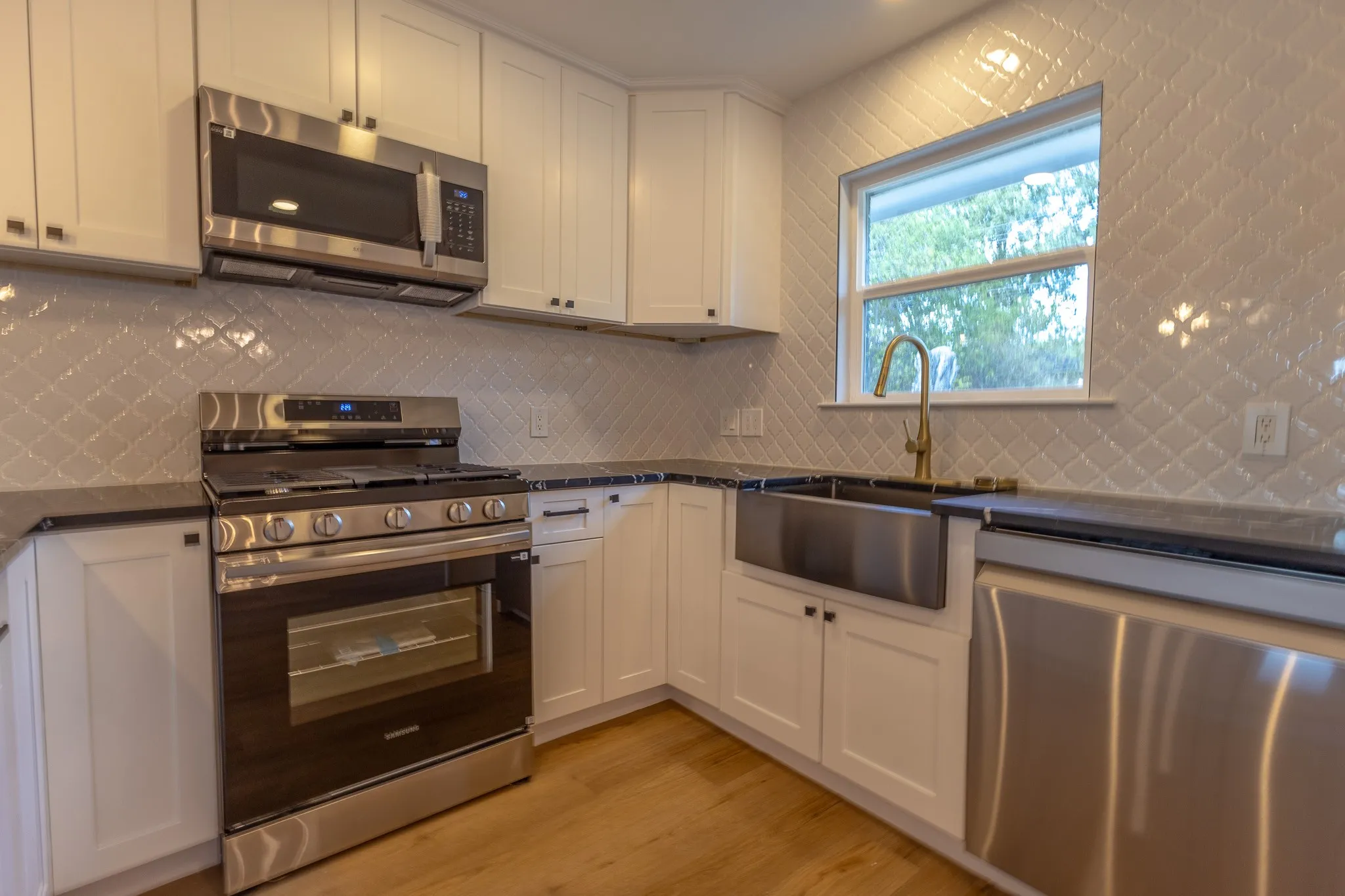 Modern kitchen featuring white shaker cabinetry, stainless steel appliances, dark countertops, a window, and wood-finish flooring