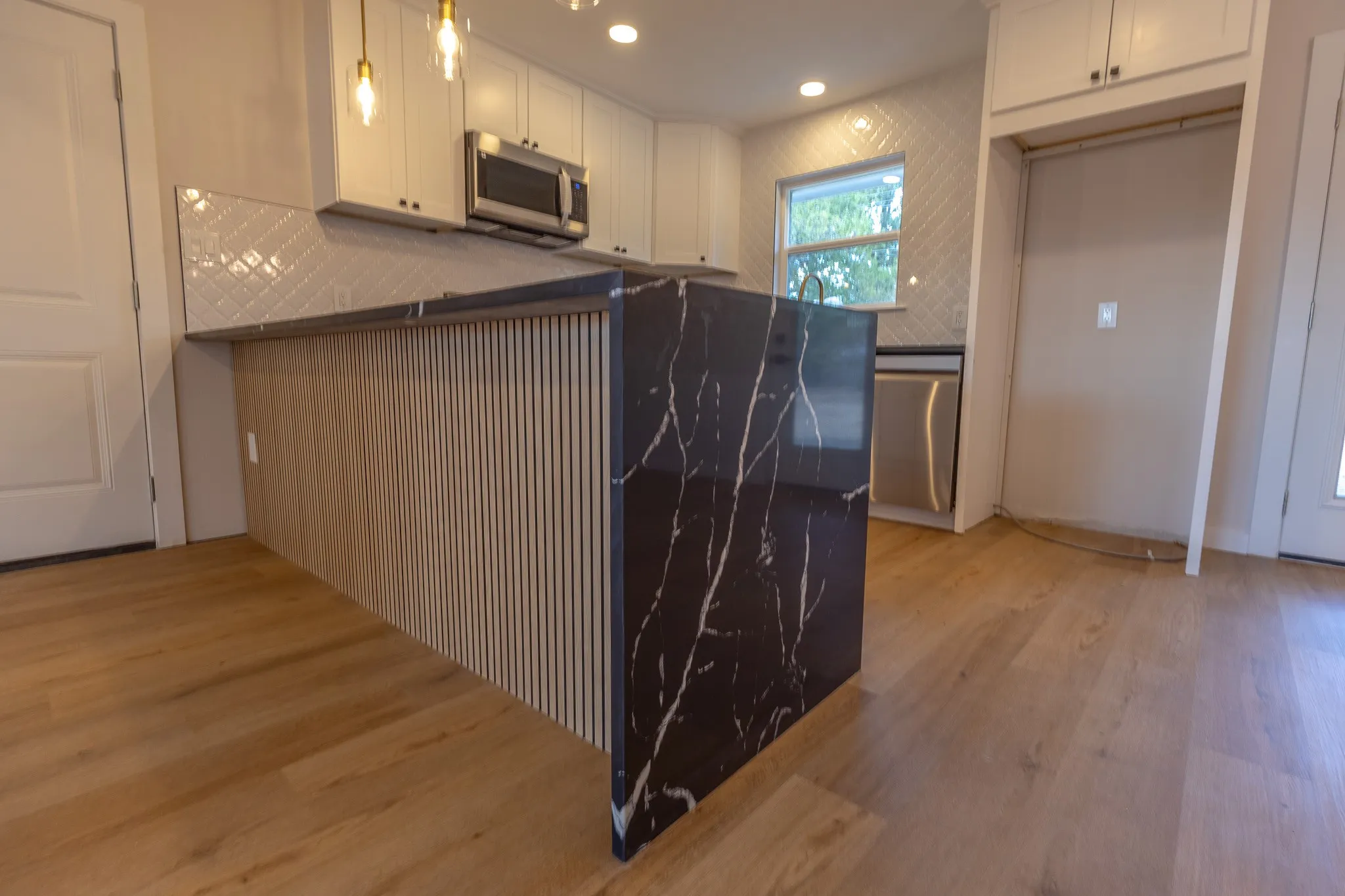 Kitchen featuring white shaker cabinetry, stainless steel microwave, black island with white veining, wood-finish flooring, and a subway tile backsplash