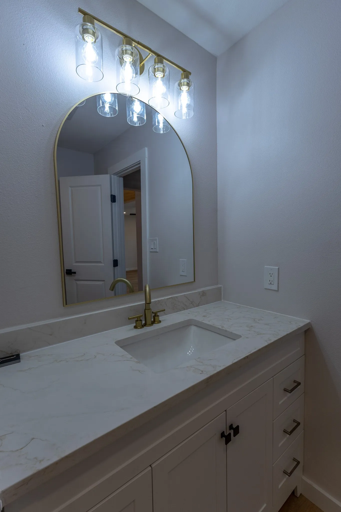 Vanity featuring a gold-framed arched mirror, a four-light fixture with clear glass shades, an integrated rectangular sink, a brass faucet, and white cabinetry with brass hardware
