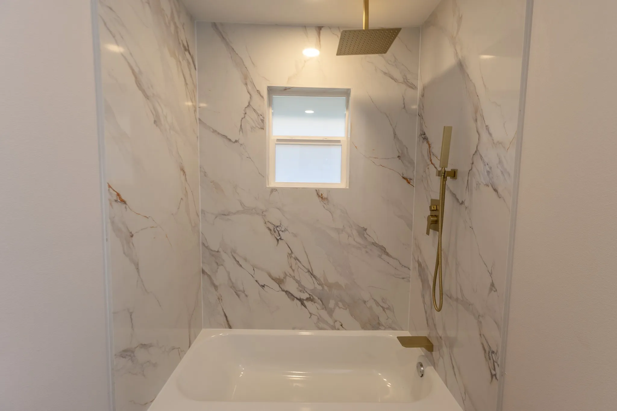 Bathtub with a window, marble-look shower surround, gold-finish rain shower head, and gold-finish handheld shower sprayer