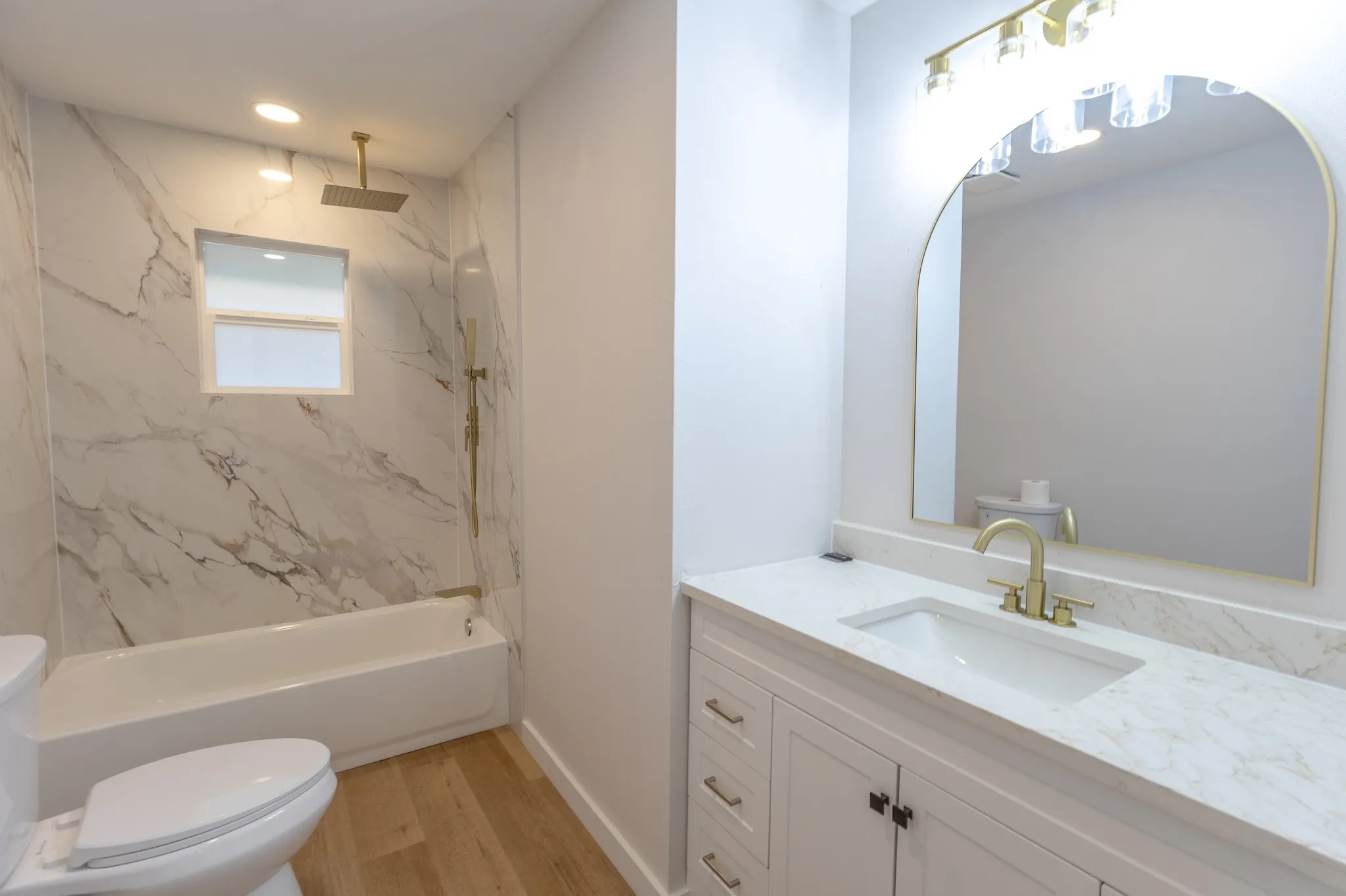 Bathroom featuring a white vanity with a light-toned countertop, a gold-framed arched mirror, and a tub/shower combination with marble-patterned wall panels