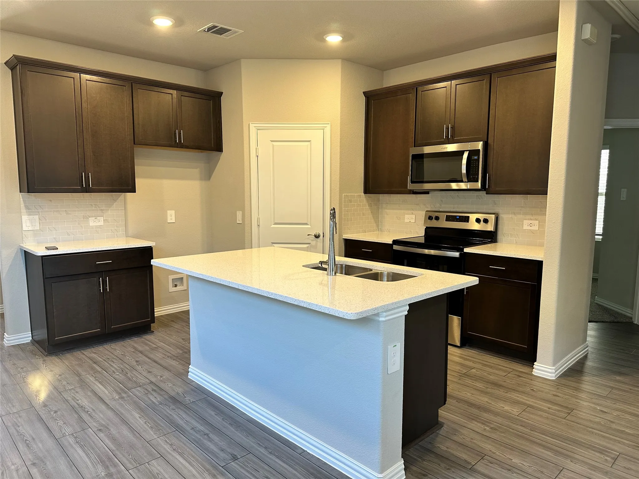 Kitchen featuring wood-finish cabinetry, stainless steel appliances, a center island with integrated sink, light-colored countertops, and a subway tile backsplash