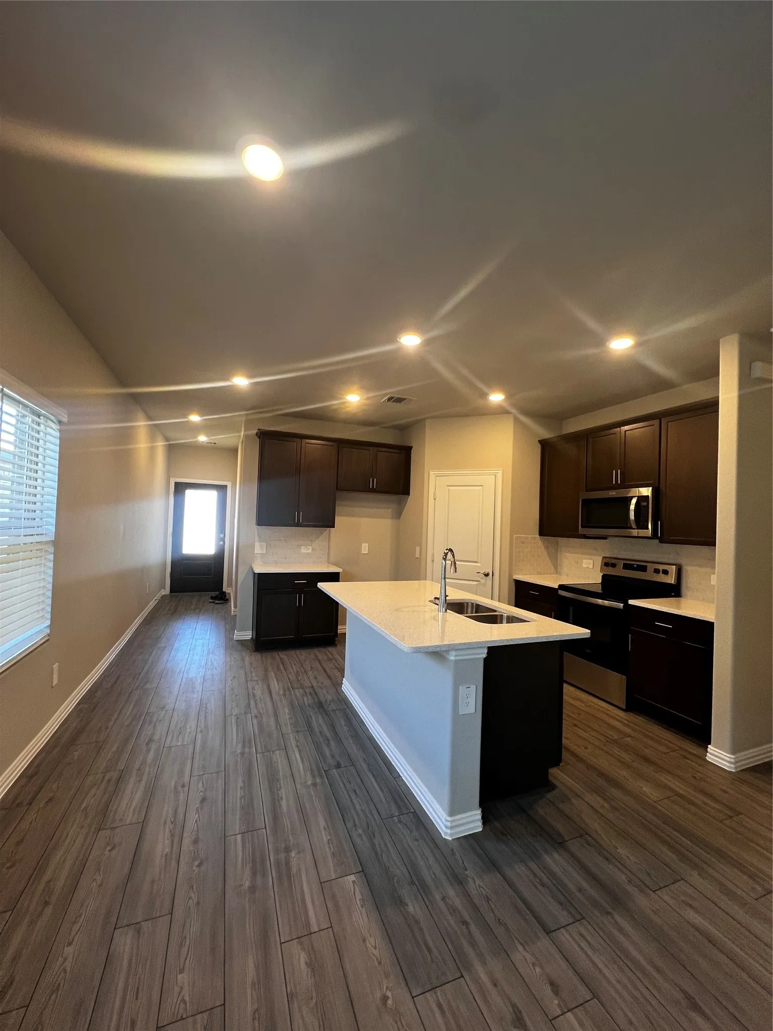 Open-concept kitchen with wood-finish flooring, recessed lighting, dark wood cabinetry, light-toned countertops, and a central island with an integrated sink