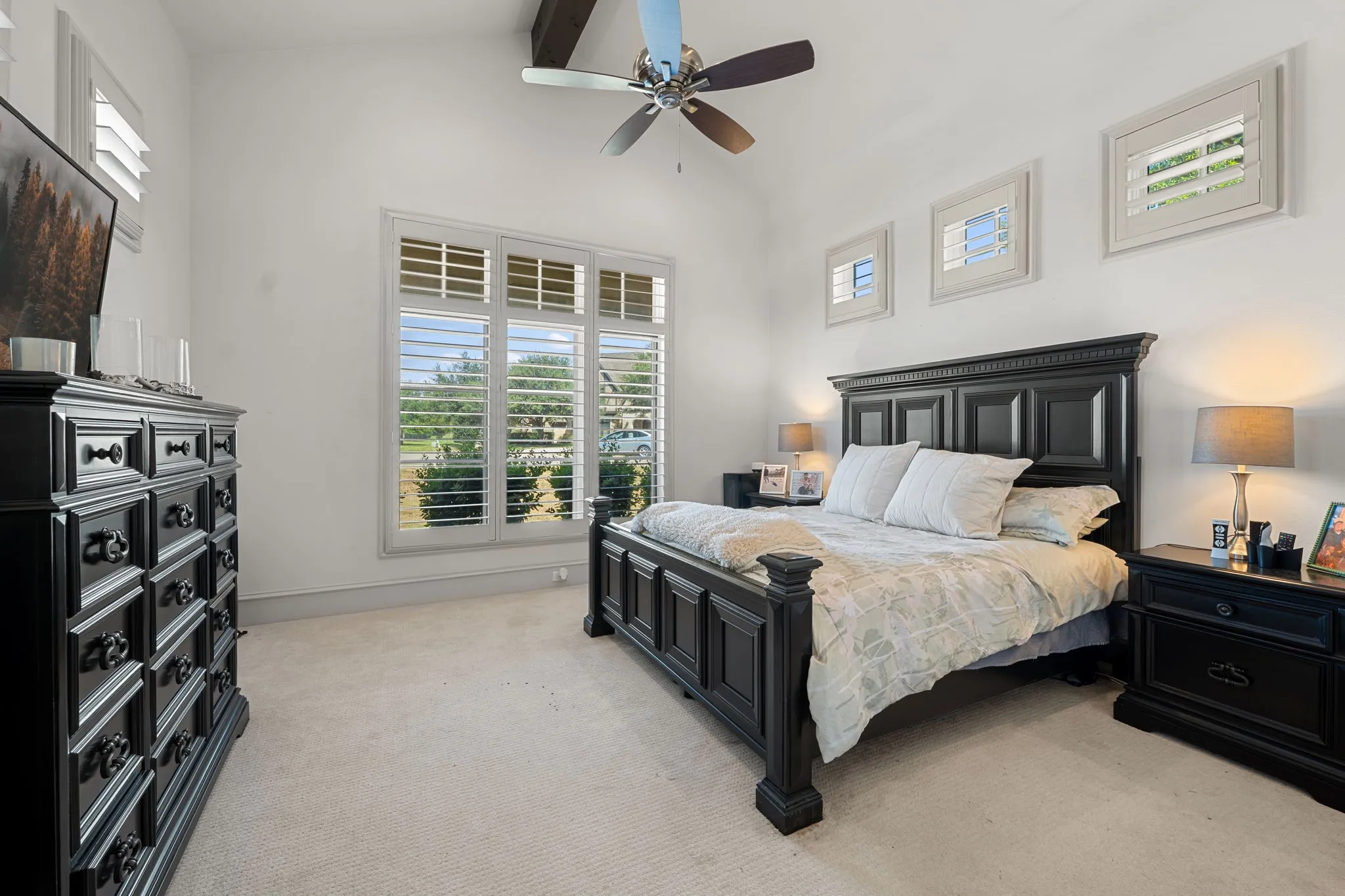 Vaulted ceiling with exposed wood beams, ceiling fan with light, large window with plantation shutters, and carpet flooring
