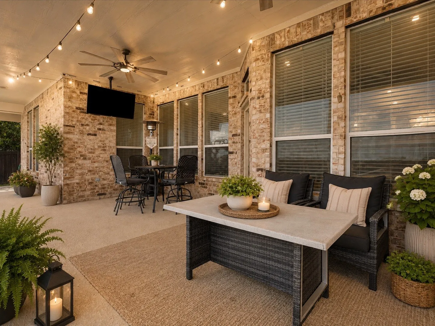 Covered patio featuring a ceiling fan, string lighting, brick exterior walls, and multiple windows with blinds