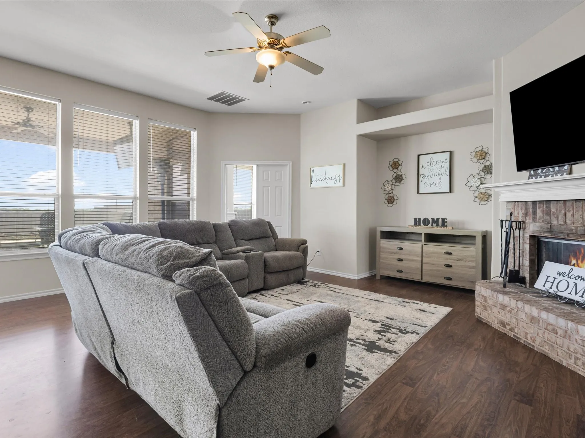 Wood-finish flooring throughout, light-colored walls, ceiling fan with integrated lighting, brick-faced fireplace with white mantel, built-in wall niche