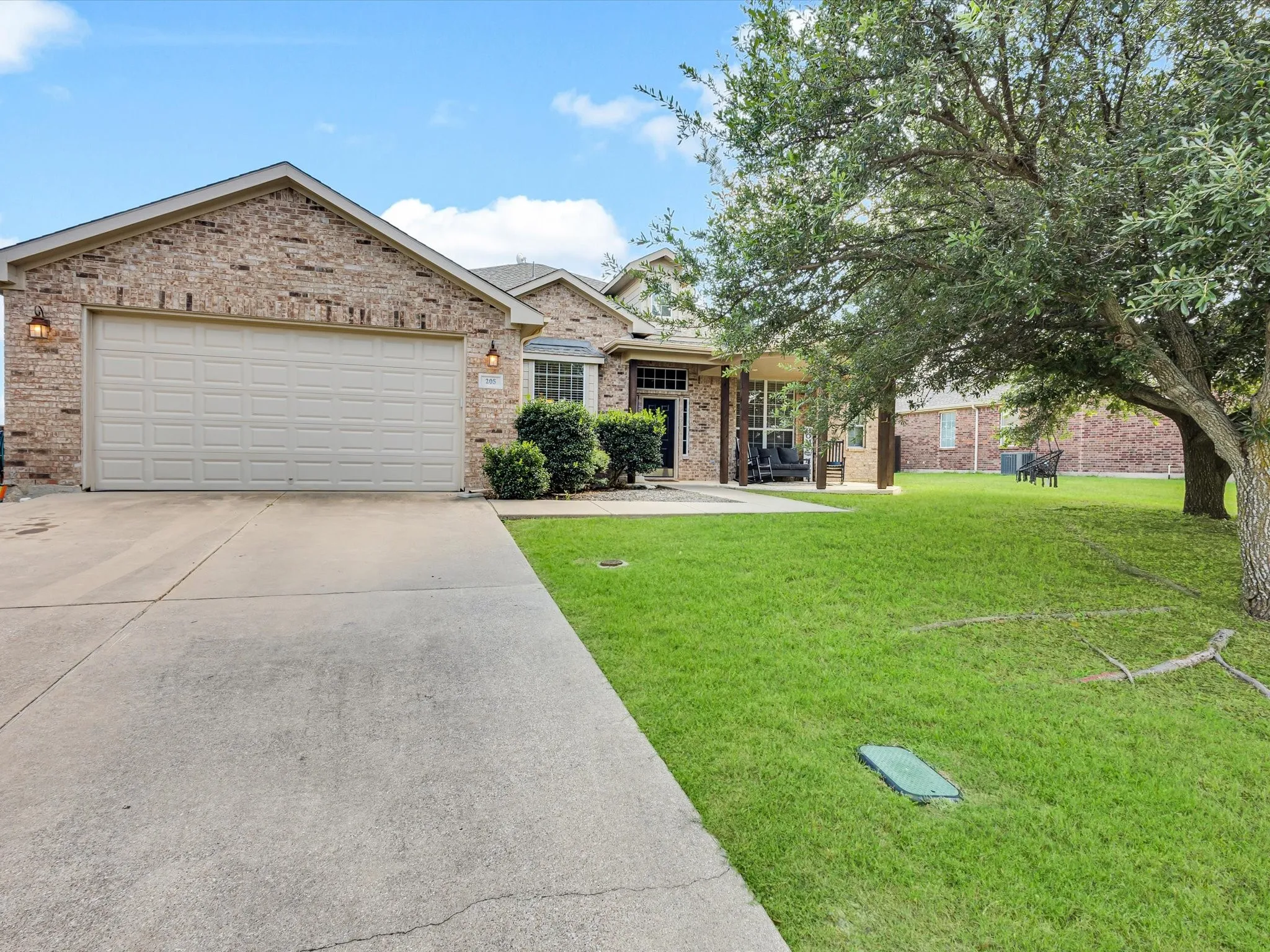 Brick exterior with covered front porch, attached garage, concrete driveway, and mature trees