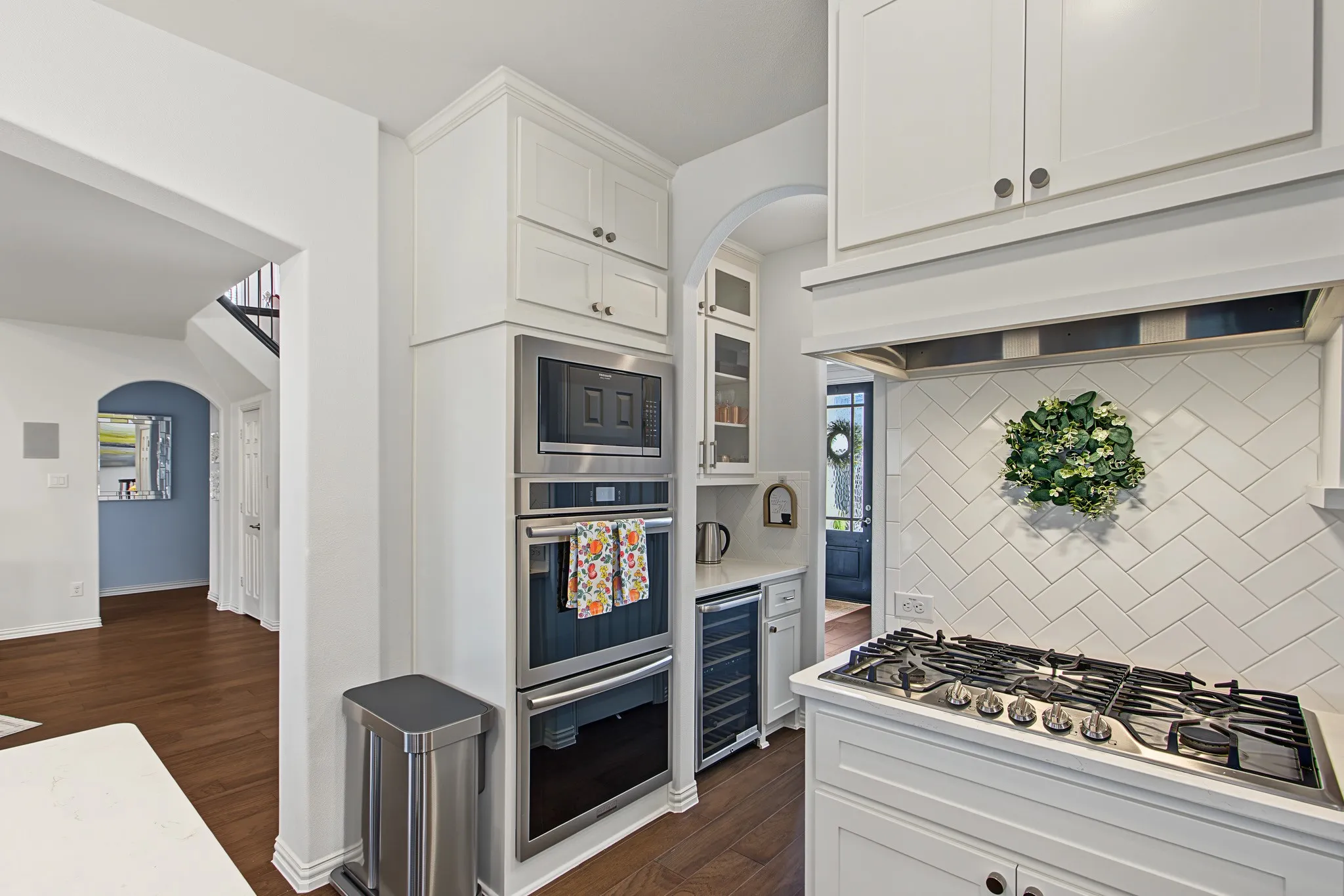 White shaker cabinetry with crown molding, stainless steel built-in double ovens, stainless steel gas cooktop, white subway tile backsplash in a herringbone pattern, and wood-finish flooring