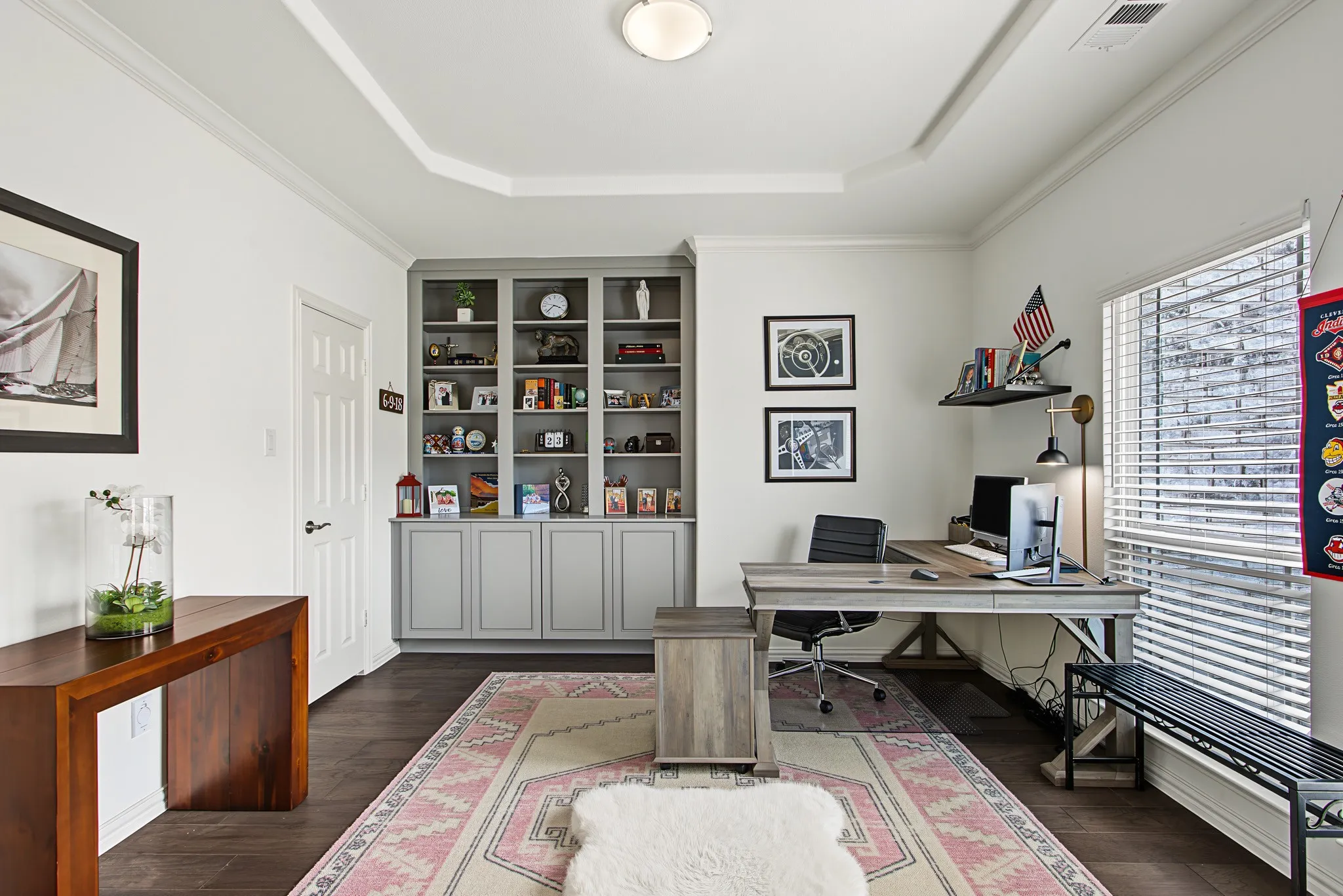 Recessed ceiling with layered crown molding, integrated built-in shelving, wood-finish flooring, and natural light from a large window