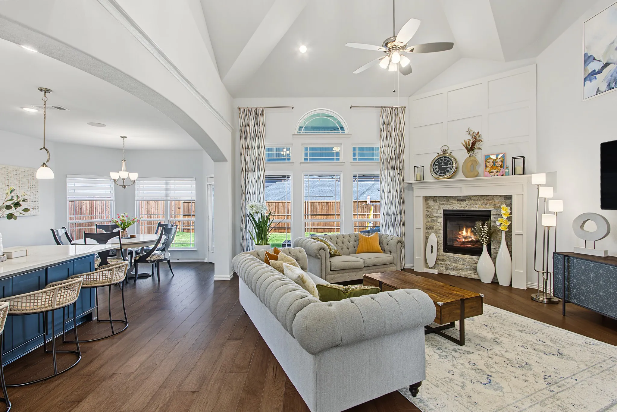 Open-concept living space featuring vaulted ceilings, wood-finish flooring, a stone-surround fireplace with a detailed mantel, and large windows with transom accents
