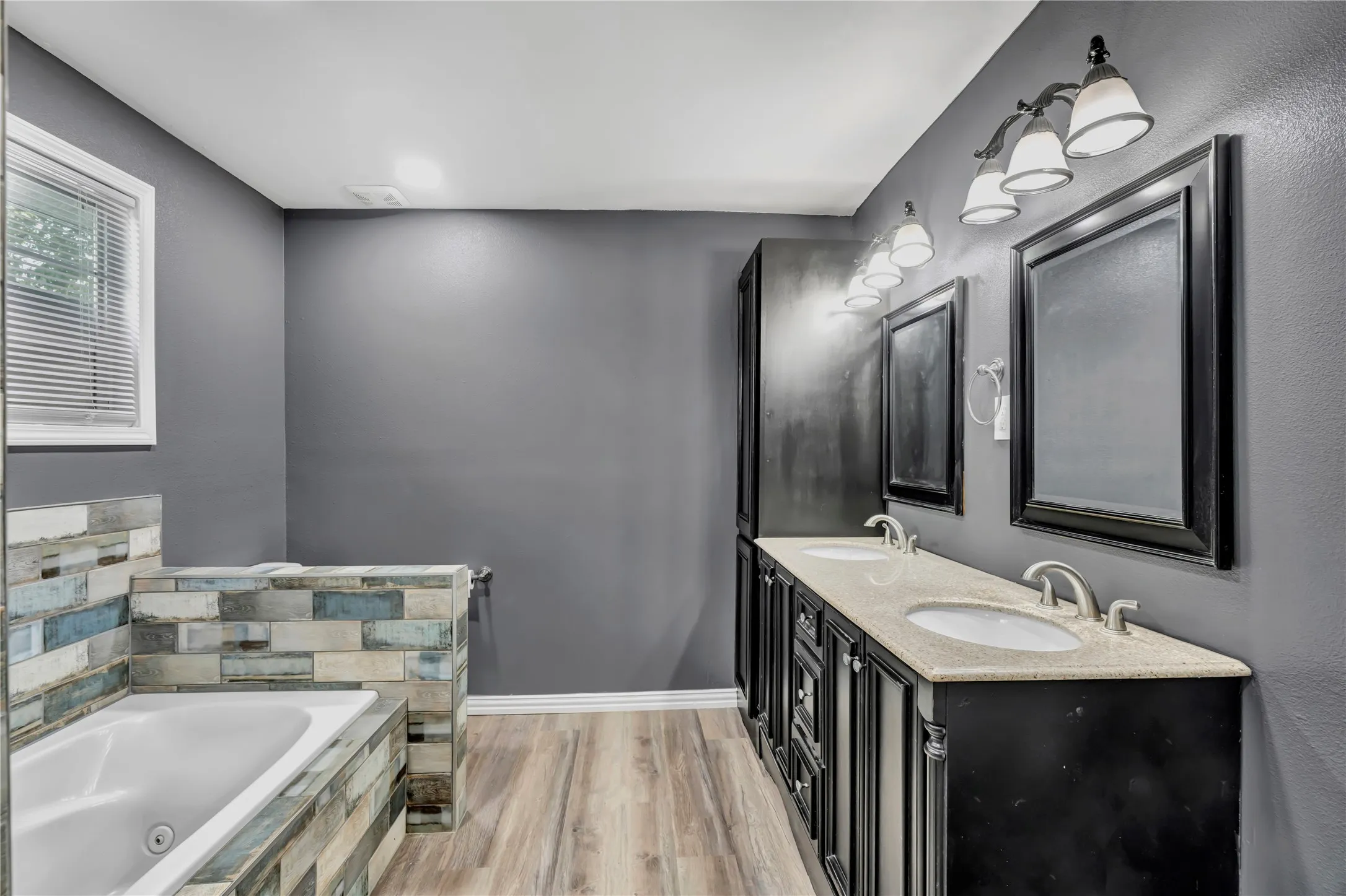 Dual vanity with stone-finish countertop and undermount sinks, wood-finish flooring, built-in soaking tub with stacked stone surround, and window with blinds