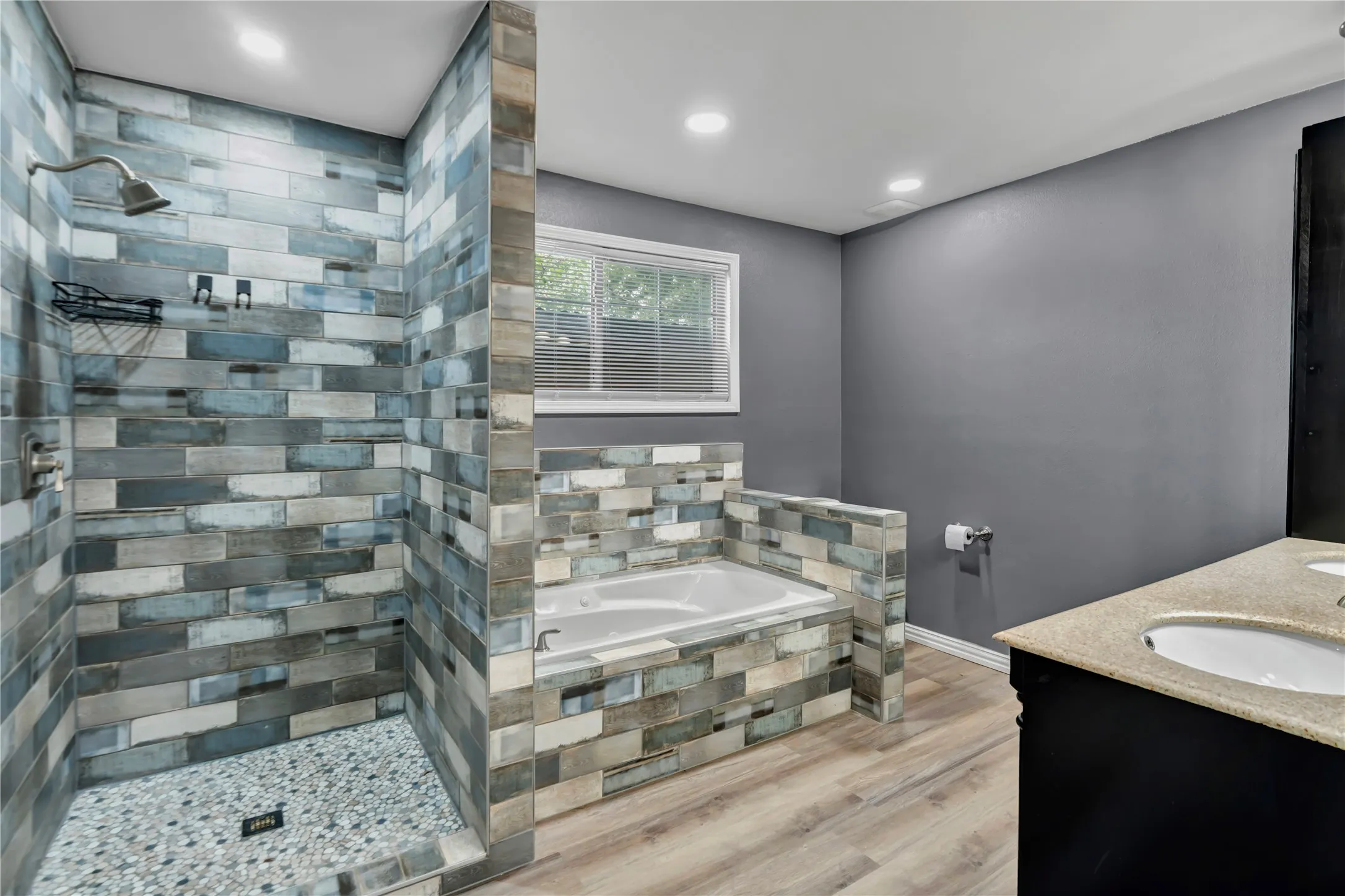 Walk-in shower with pebble tile floor, built-in soaking tub, dual vanity with undermount sinks, and wood-finish flooring