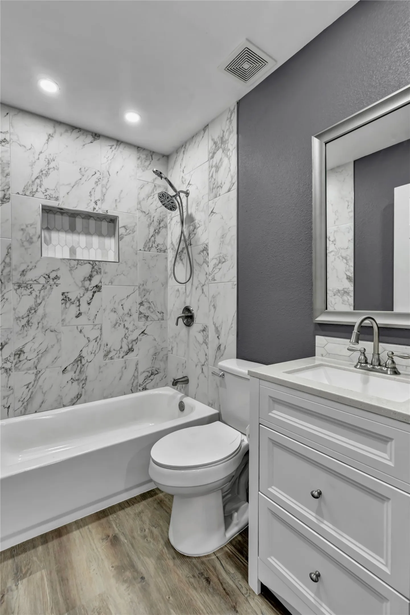 Marble-look tile shower surround with a built-in niche, white bathtub, wood-finish flooring, and a white vanity with a countertop
