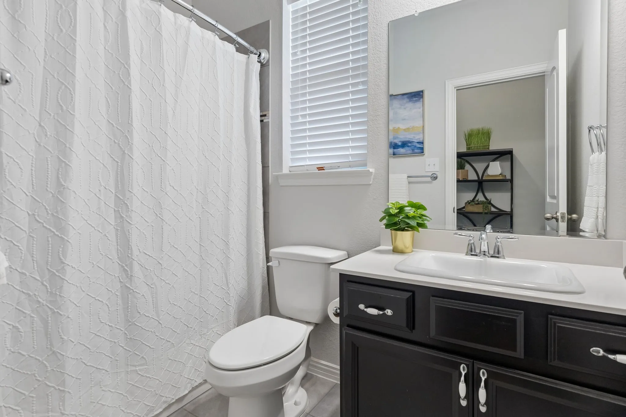 White shower curtain with subtle pattern, white toilet, dark wood-finish vanity with white countertop, chrome faucet, and rectangular mirror