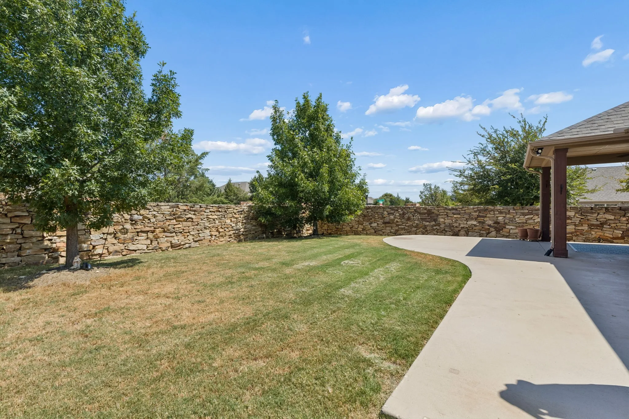 Expansive backyard featuring a stone privacy wall, mature trees, a manicured lawn, and a covered patio with a curved concrete walkway