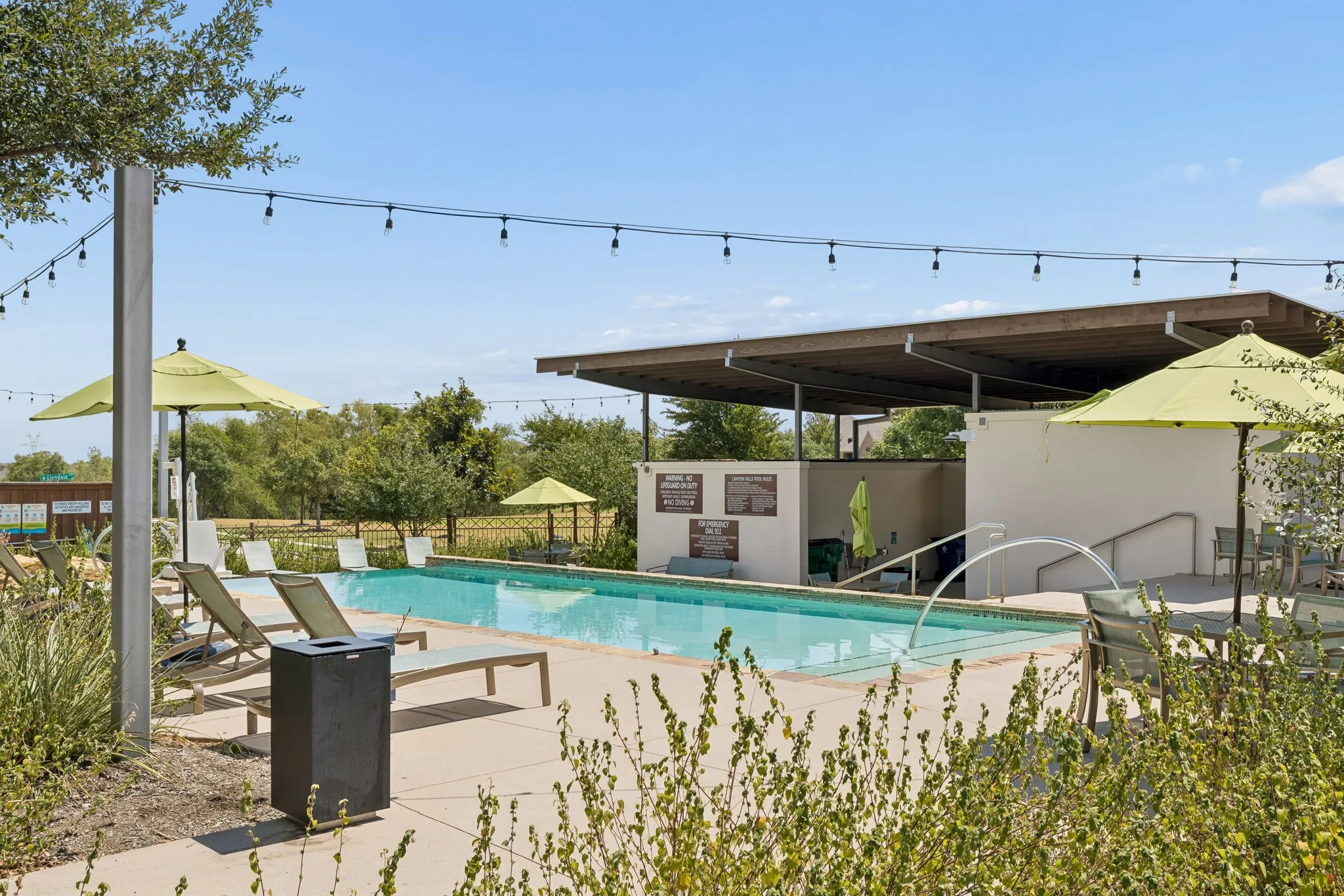 Outdoor swimming pool with integrated water feature, adjacent concrete deck, string lighting, and covered amenity structure