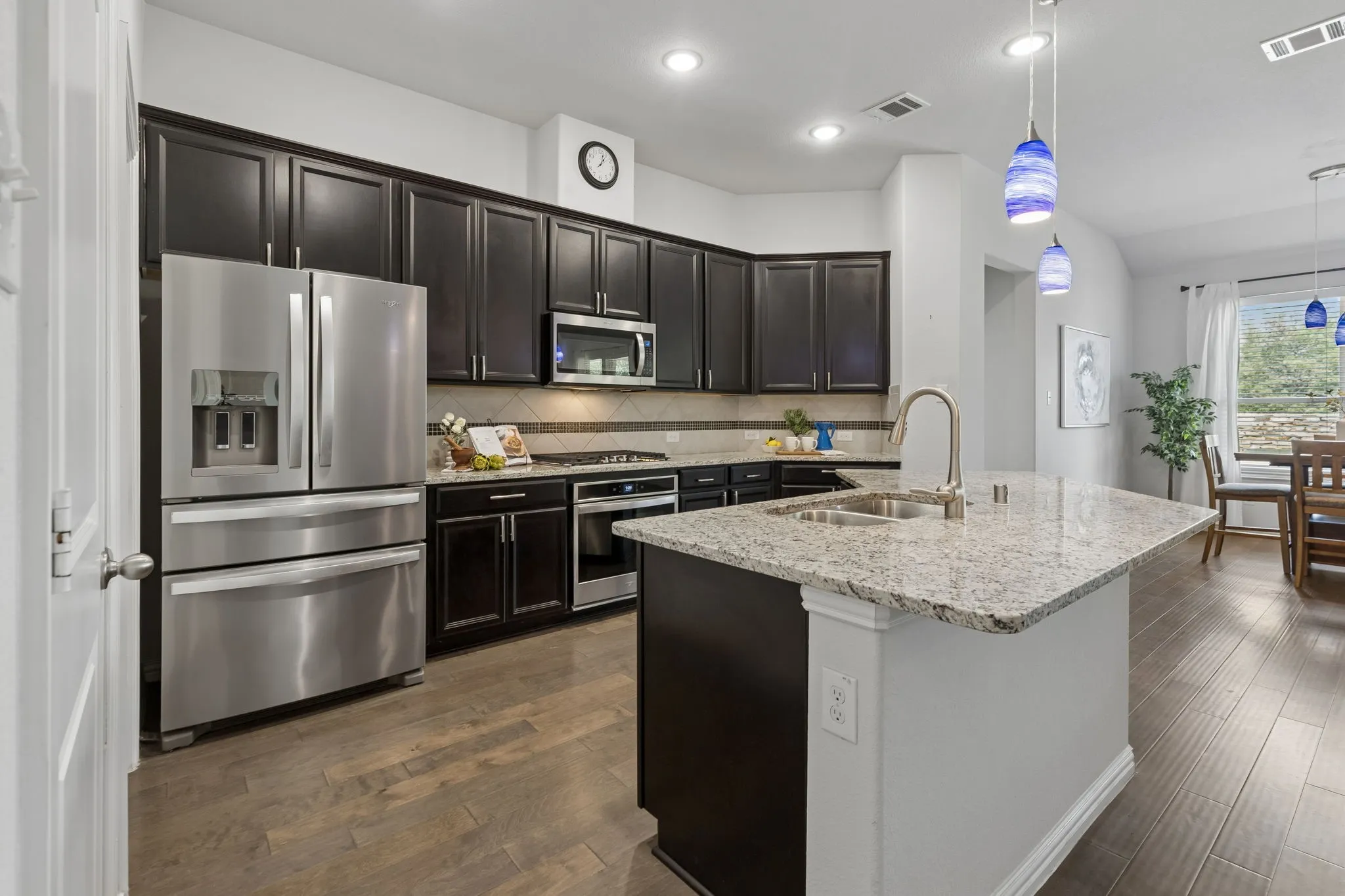 Kitchen featuring dark wood cabinetry, stainless steel appliances, a light-toned granite island, wood-finish flooring, and pendant lighting