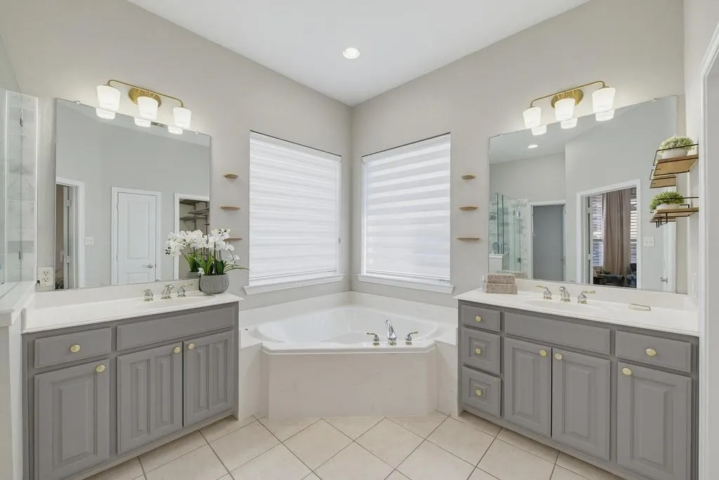 Corner soaking tub, dual gray vanity cabinets with gold hardware, two large wall-mounted mirrors, and light tile flooring