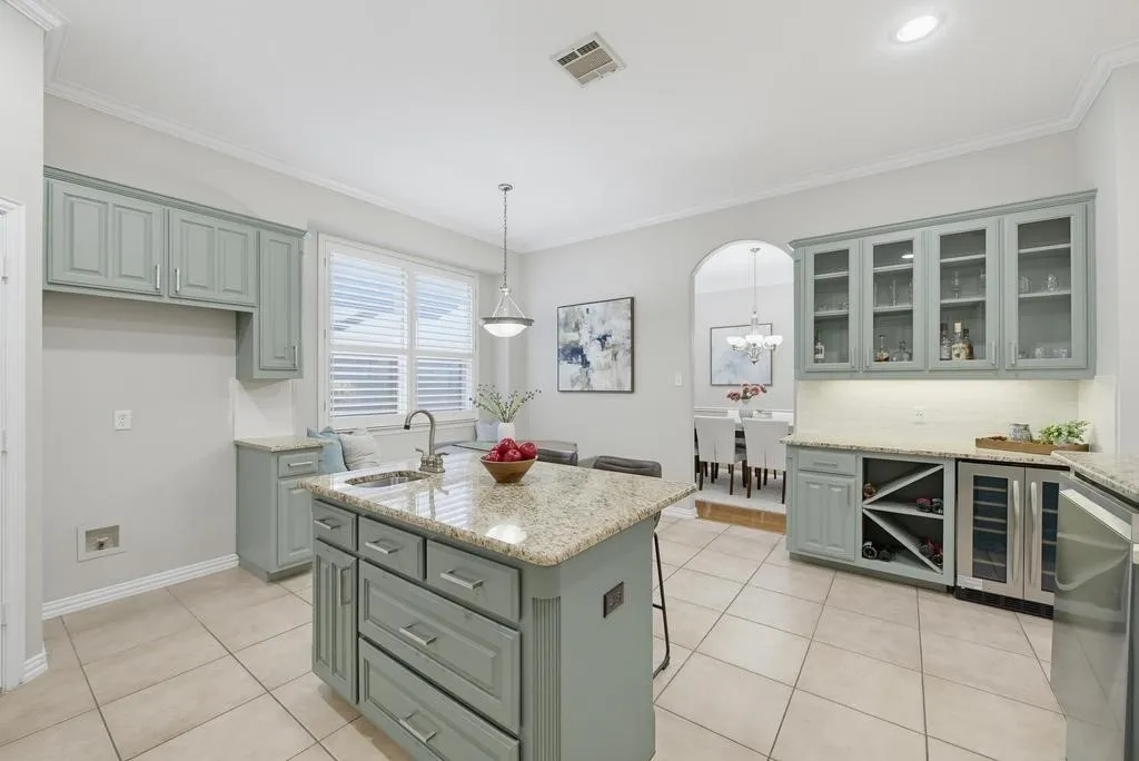 Kitchen featuring a central island with an undermount sink and stone countertop, light-toned cabinetry, tile flooring, and a built-in wine rack with a beverage refrigerator