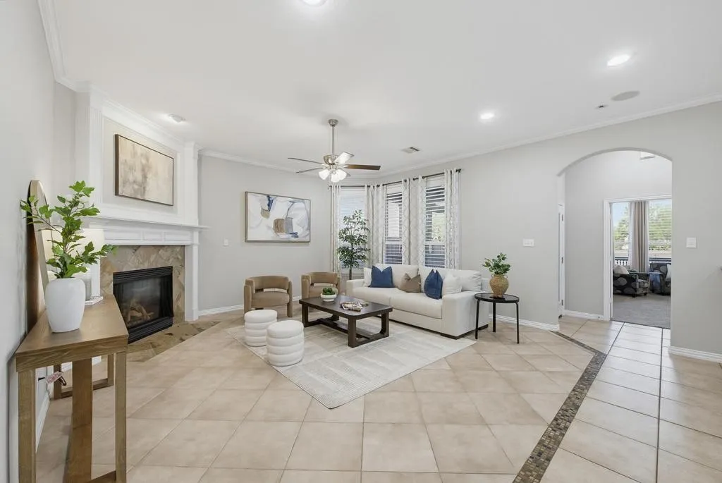 Living room featuring a fireplace with a stone surround, crown molding, a ceiling fan, tiled flooring, and an arched entryway