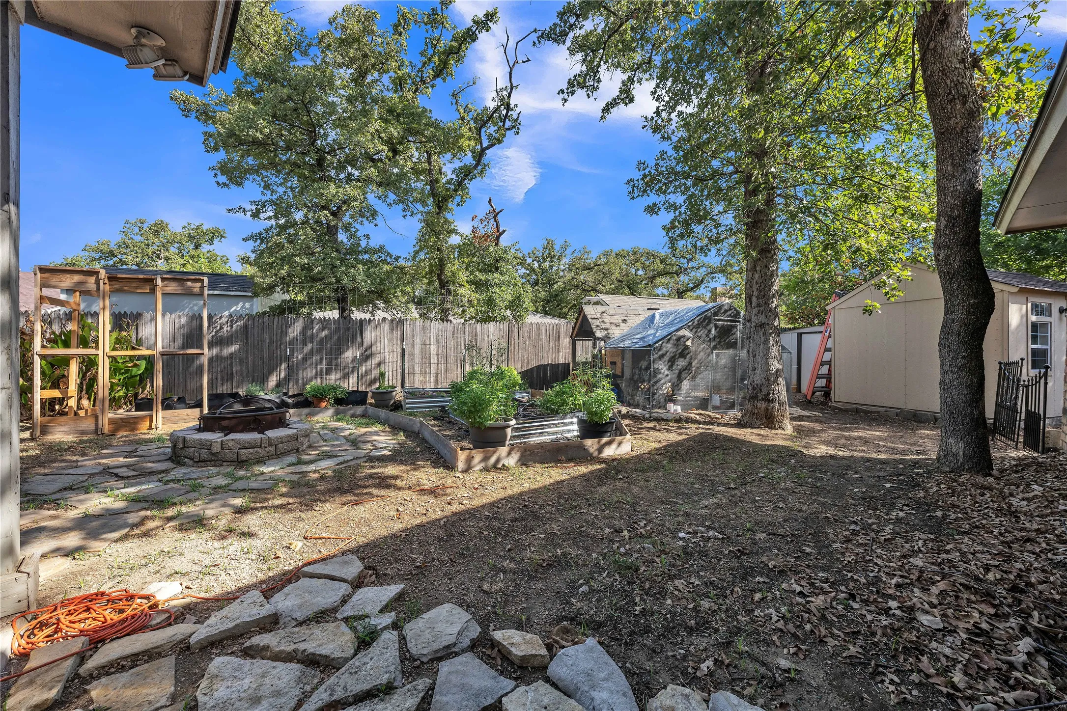 Expansive backyard featuring a stone fire pit, raised garden beds, a wood privacy fence, and mature trees