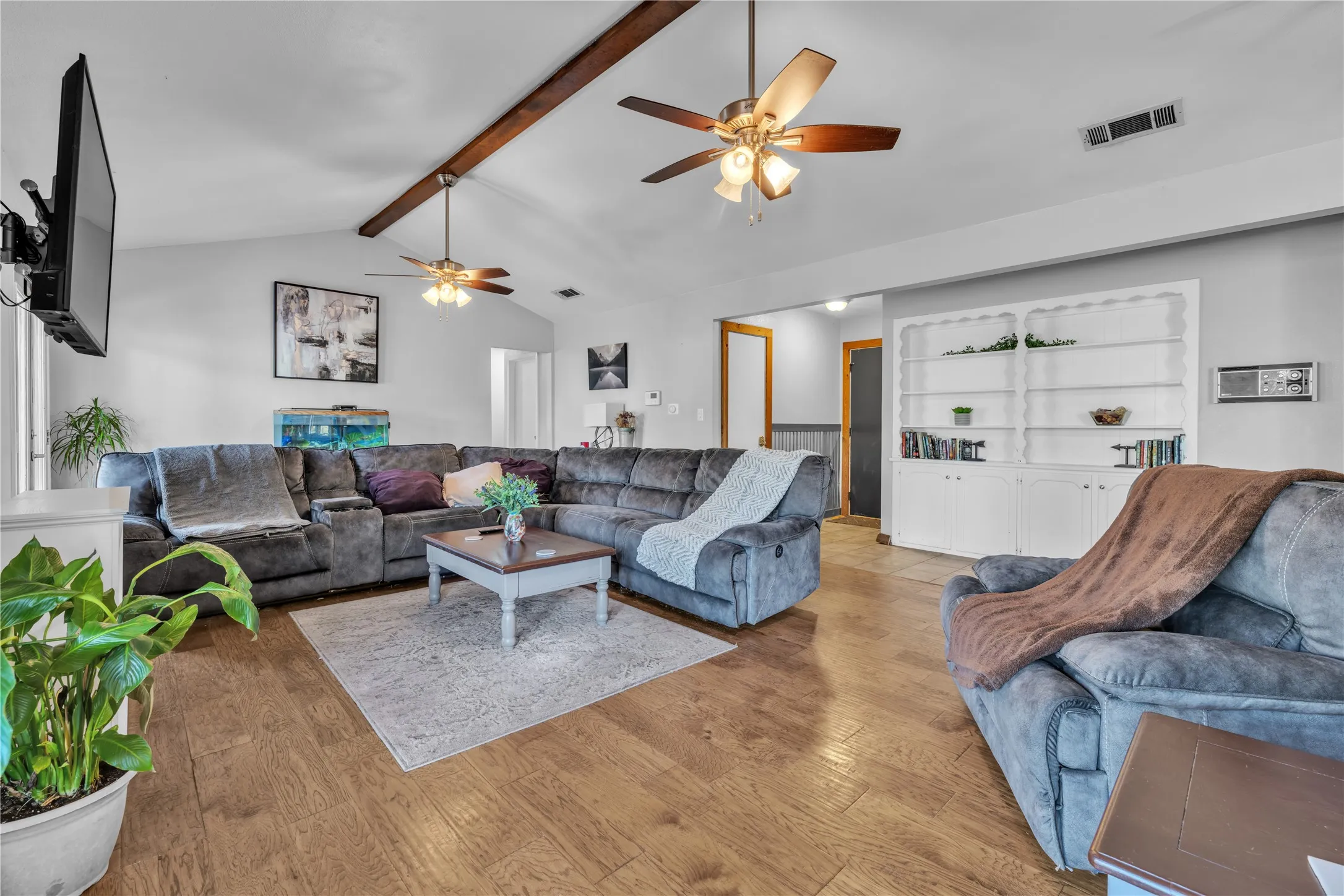 Vaulted ceiling with exposed wood beams, wood-finish flooring, two ceiling fans with light kits, and built-in shelving with lower cabinetry
