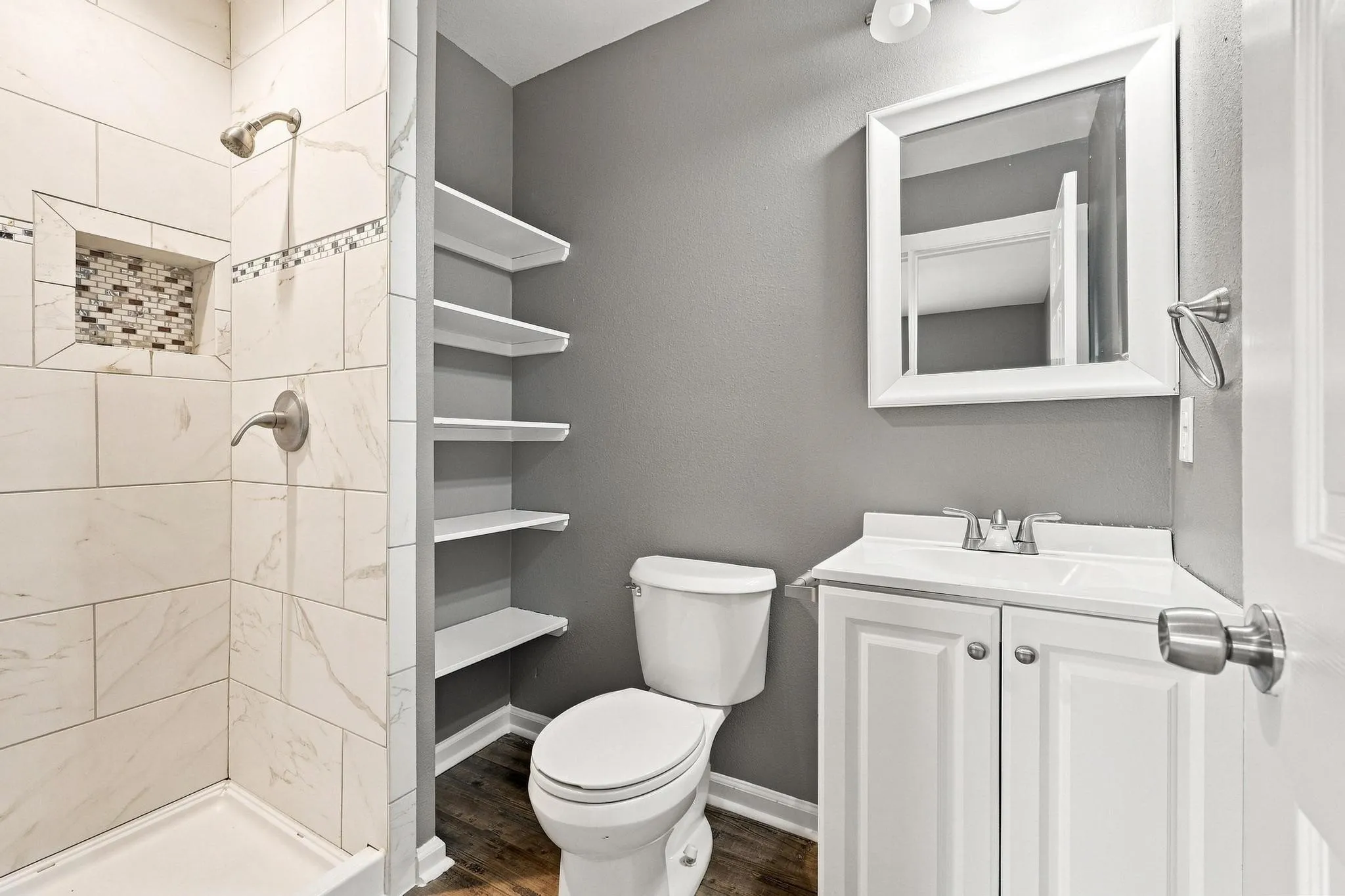 Tiled shower with mosaic accent, built-in shelving, wood-finish flooring, and a white vanity with mirror