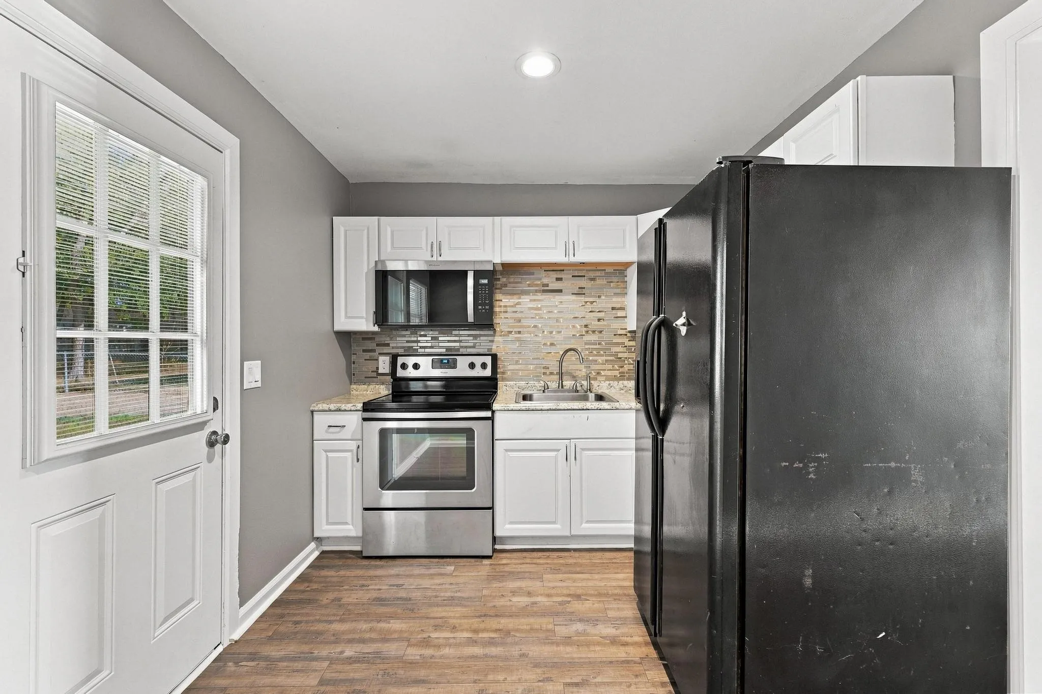White cabinetry, stainless steel range and microwave, light-toned backsplash, granite-finish countertops, and wood-finish flooring