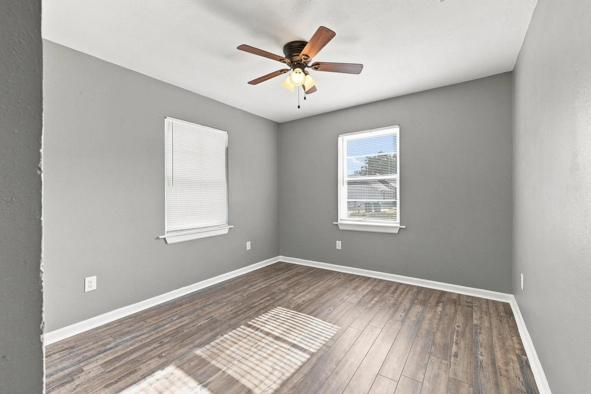Wood-finish flooring, light grey painted walls, white trim, two windows, and a ceiling fan with integrated lighting
