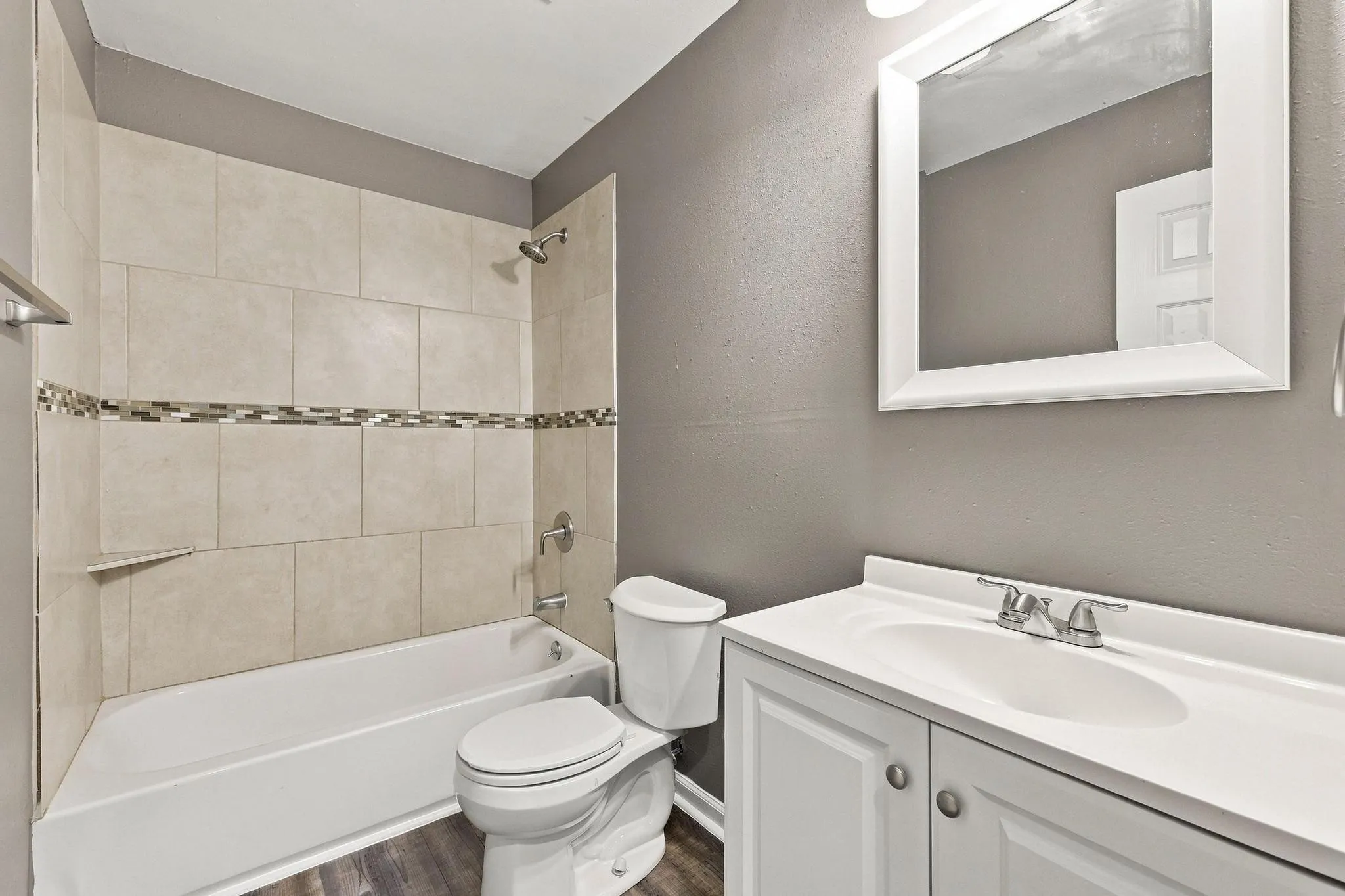 Bathroom featuring a white bathtub, tiled shower surround with decorative accent, white vanity with integrated sink, framed mirror, and wood-finish flooring
