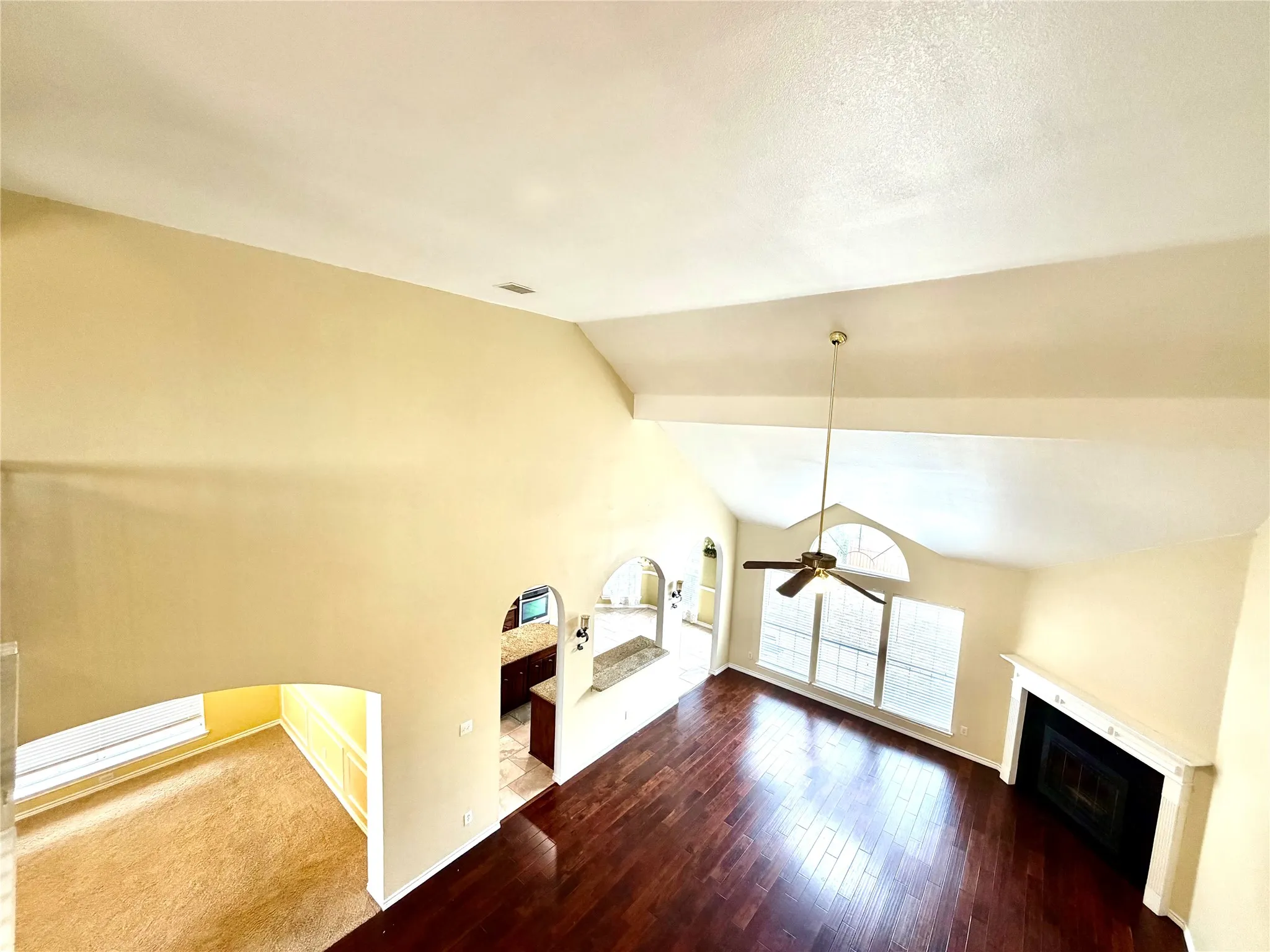 Vaulted ceiling with a ceiling fan, wood-finish flooring, arched doorways, a fireplace with a white mantel, and large windows