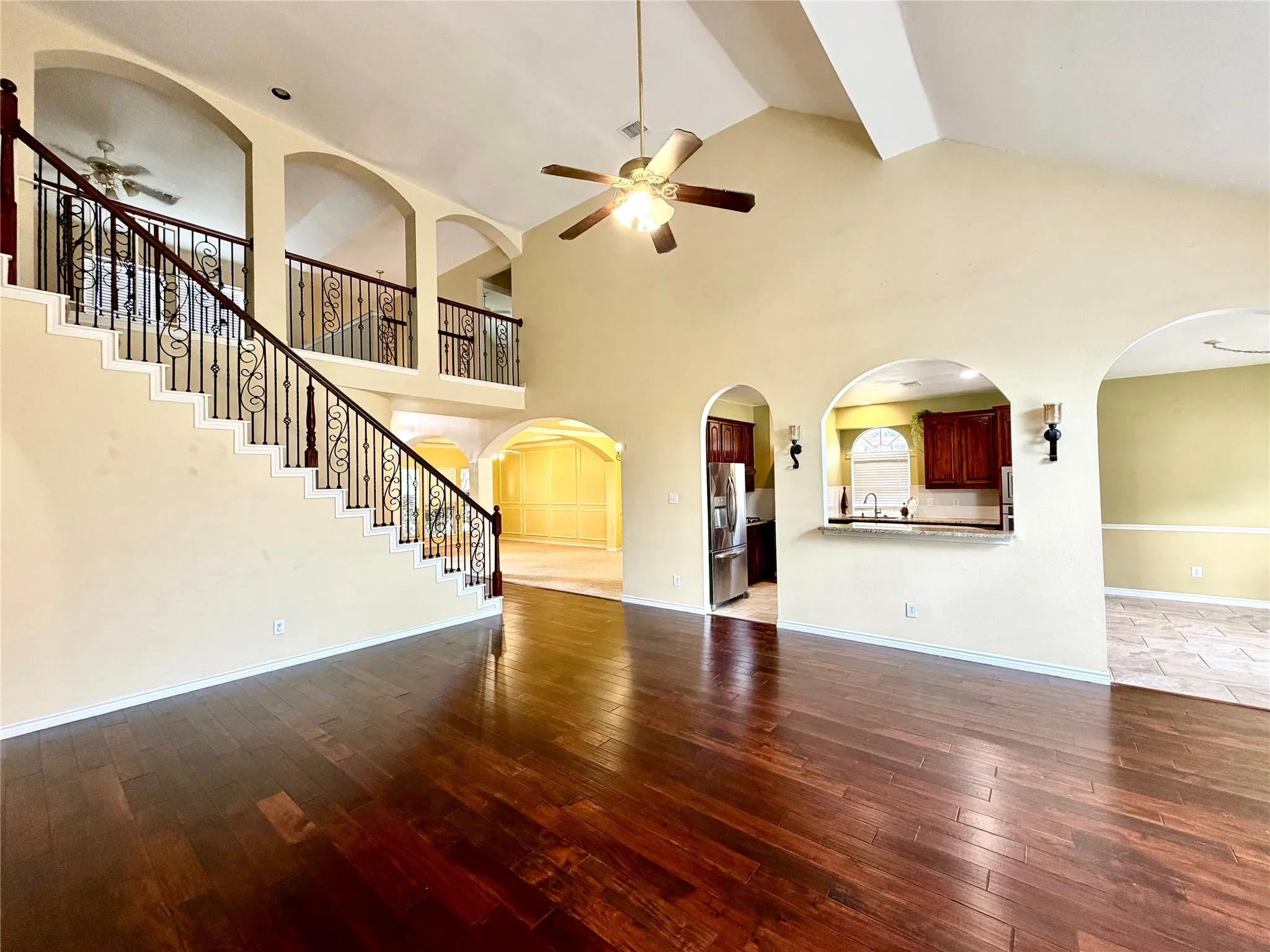 Vaulted ceiling, dark wood-finish flooring, wrought iron staircase with wood handrail, arched wall openings, and a ceiling fan with integrated lighting