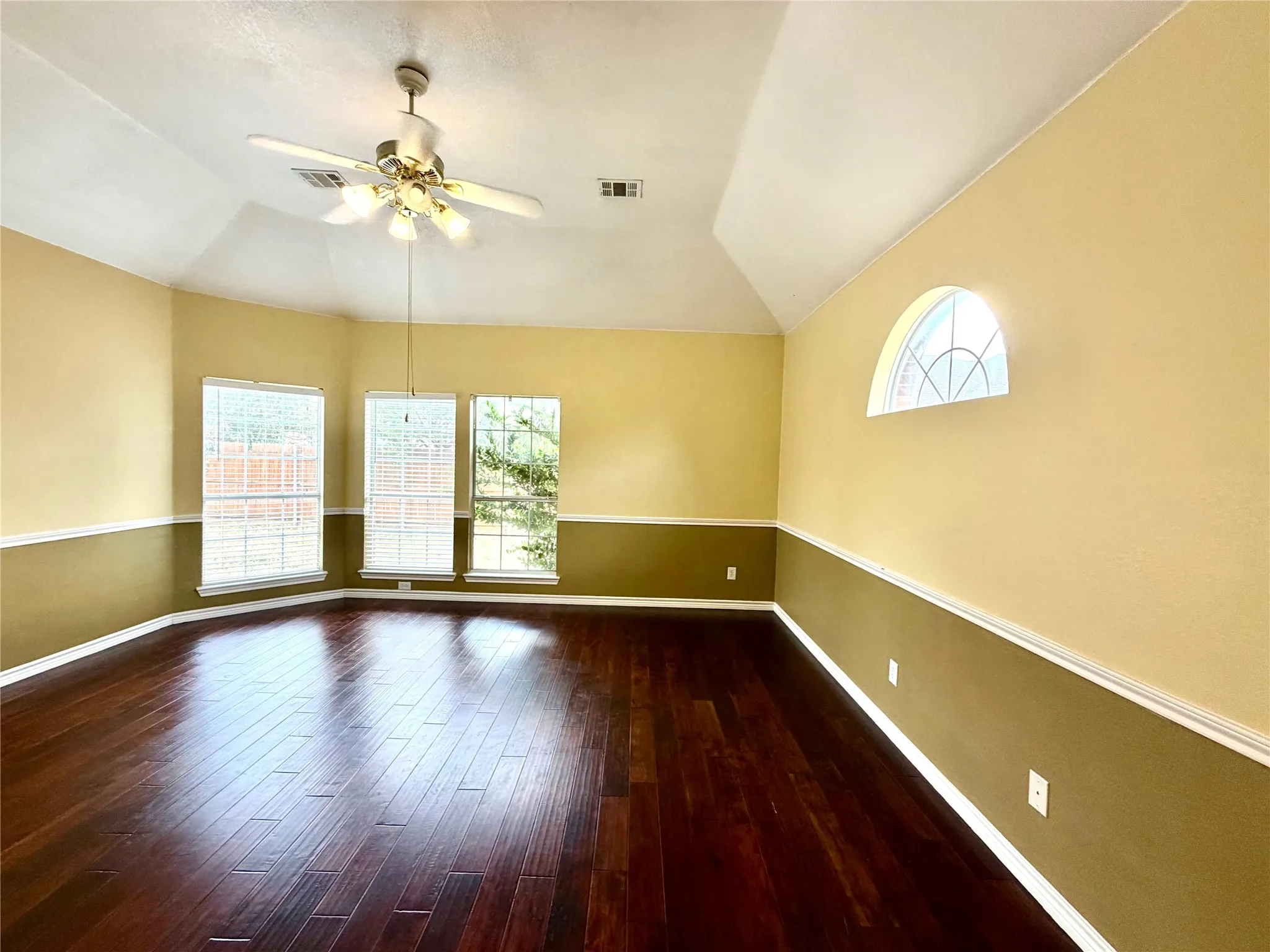Vaulted ceiling, dark wood-finish flooring, architectural arched window, three distinct windows, and a ceiling fan with integrated lighting
