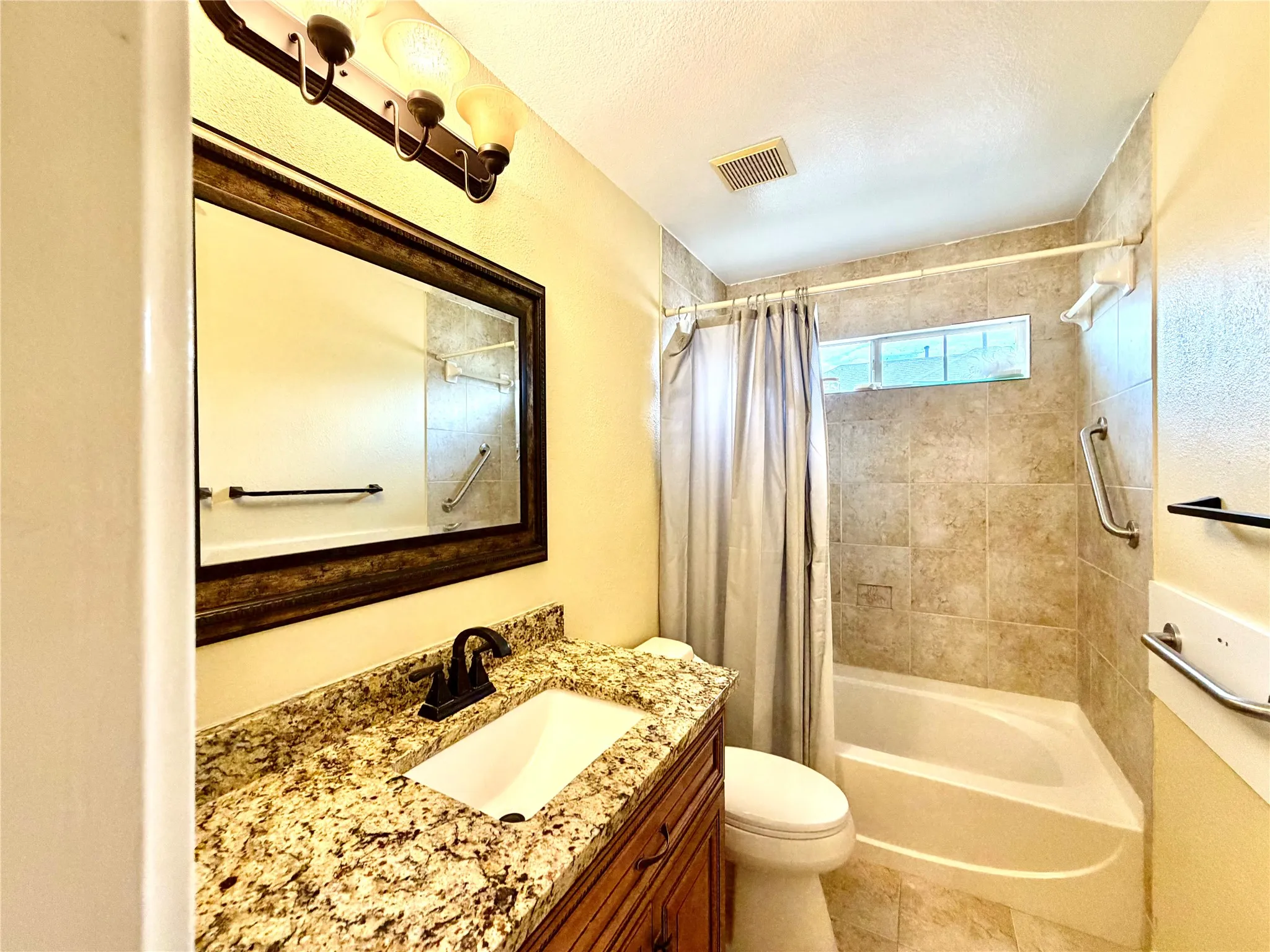 Bathroom featuring a wood-finish vanity, stone-finish countertop, undermount sink, a framed mirror, and a tiled shower-tub combination with grab bars