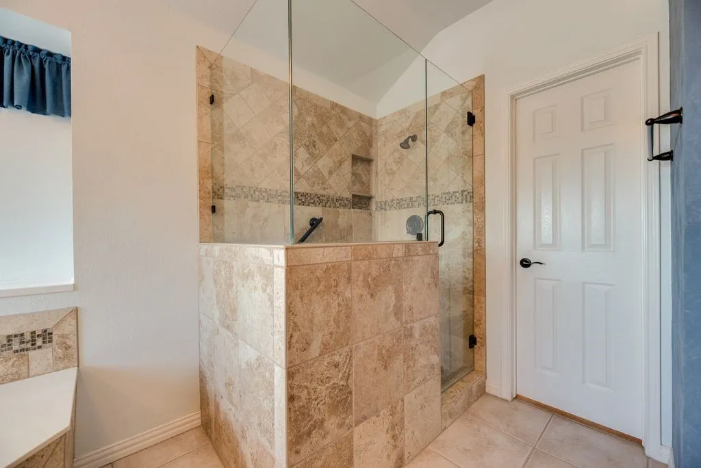 Tiled shower enclosure with a clear glass door, built-in shelving, and a decorative tile accent band