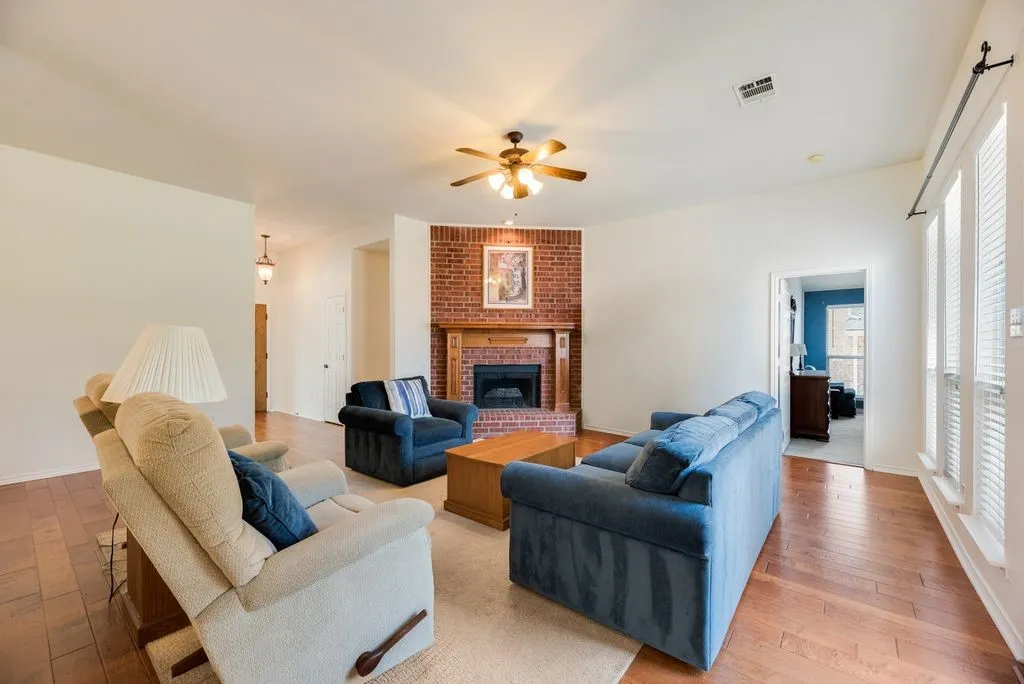 Brick fireplace with wood mantle, wood-finish flooring, ceiling fan, and white window blinds