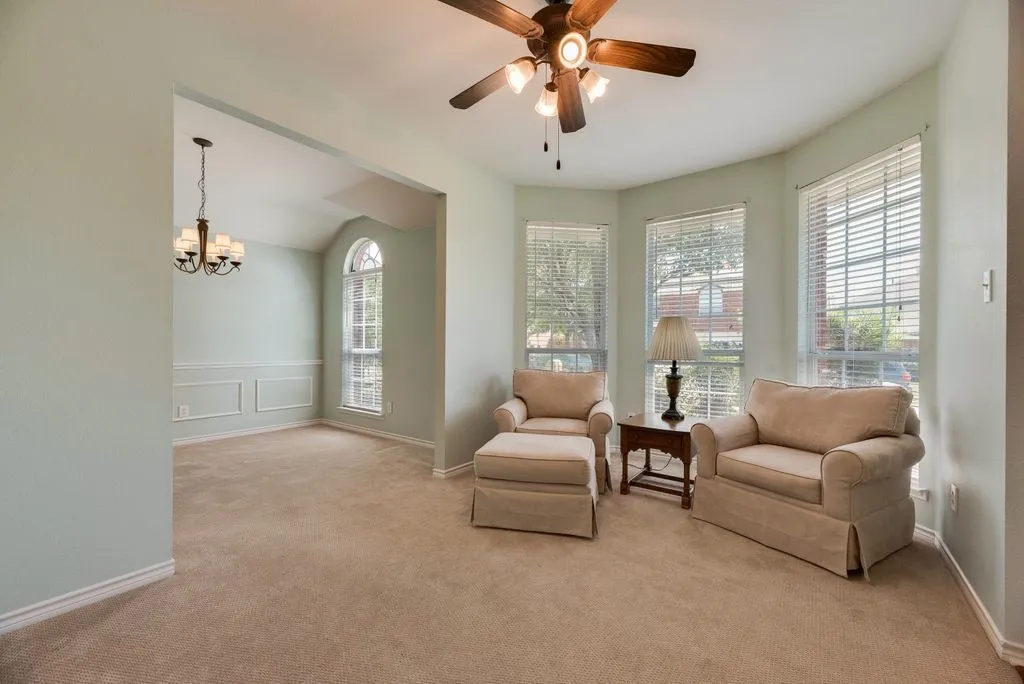 Carpeted living area featuring a ceiling fan, bay windows with blinds, and an arched entry to a room with wainscoting and a chandelier