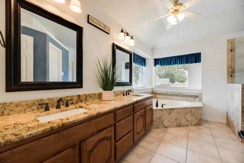 Dual vanity with granite countertops, wood cabinetry, built-in soaking tub with tile surround, separate tiled shower, and vaulted ceiling with ceiling fan