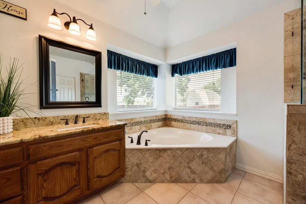 Dual vanity with granite countertop, corner soaking tub with tile surround, tile flooring, and vaulted ceiling