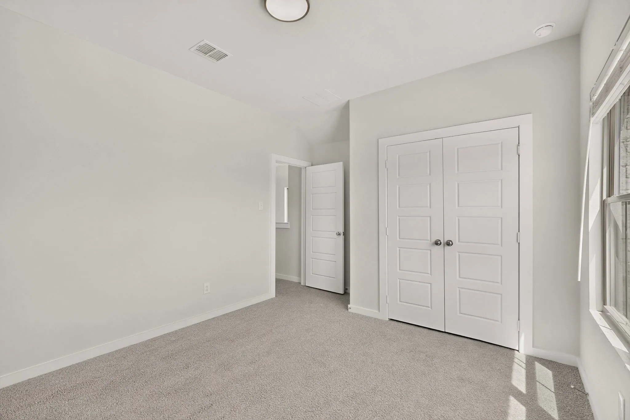 Neutral wall paint, light gray carpet, paneled bifold doors, window with white trim, and a ceiling-mounted light fixture