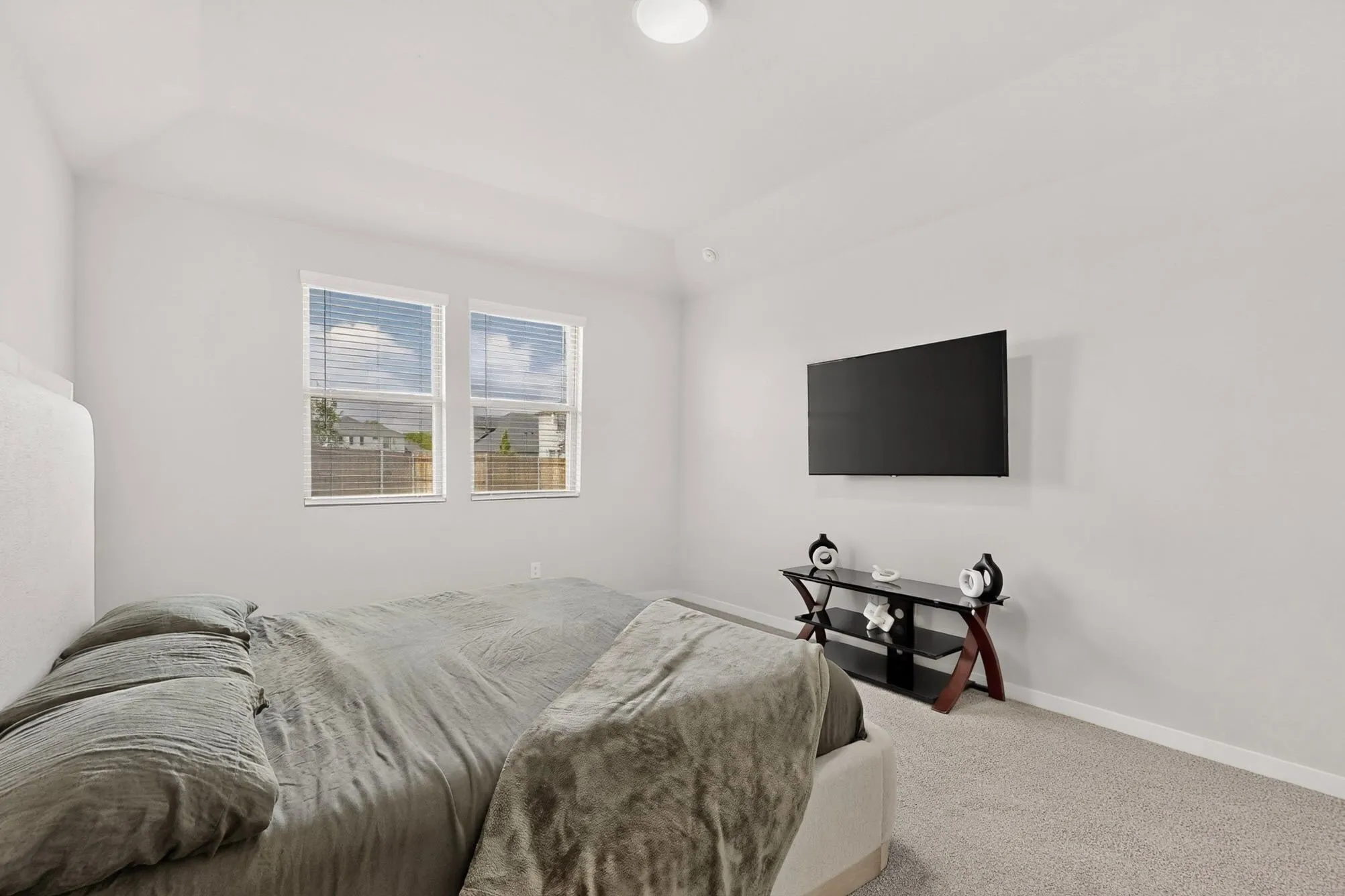 Bright secondary bedroom with dual windows featuring horizontal blinds, neutral carpeting, wall-mounted flat-screen television with black media console, and clean white walls throughout